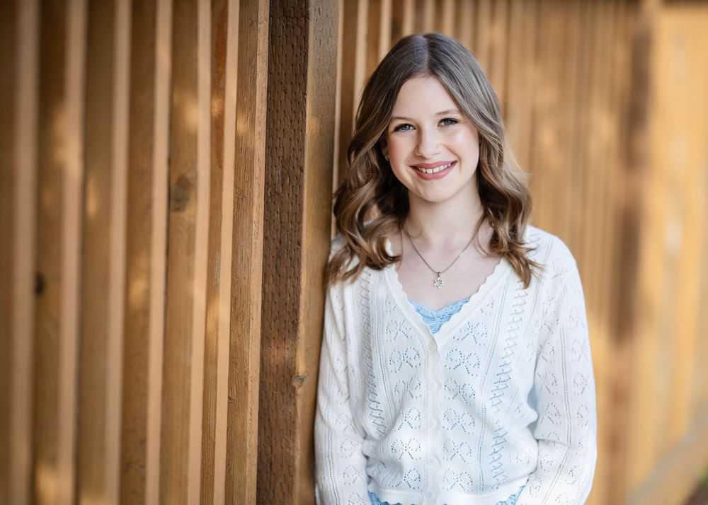 Smiling girl with long wavy brown hair wearing a white knit cardigan and necklace, leaning against a rustic wooden fence outdoors.