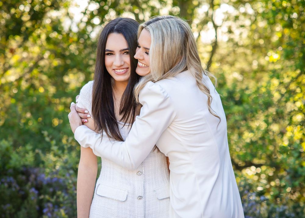 Two smiling women in matching white outfits embrace outdoors, surrounded by sunlit green foliage.