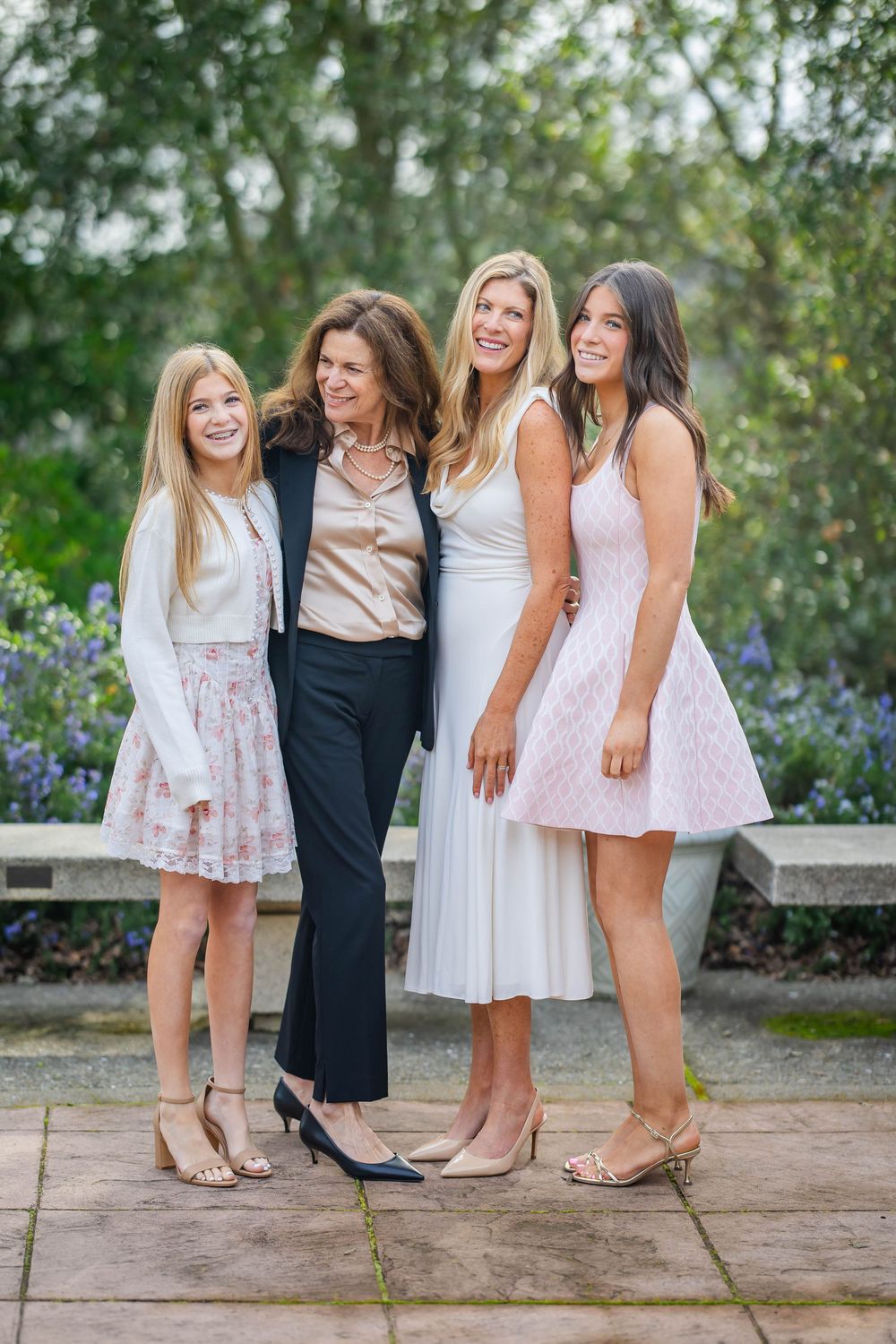 Four women of different ages in dresses and a blazer pose together smiling and arm in arm on a stone patio in a garden.