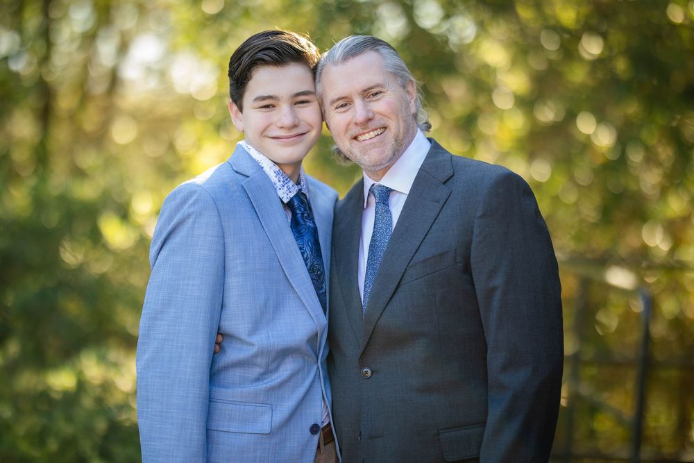 Smiling teenage boy in a light blue suit stands beside an older man in a gray suit and tie against a sunlit, leafy green background.