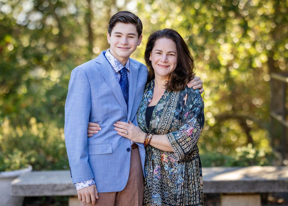 A smiling teenage boy in a light blue blazer and tie poses with his arm around a woman in a patterned dress outdoors among green trees.
