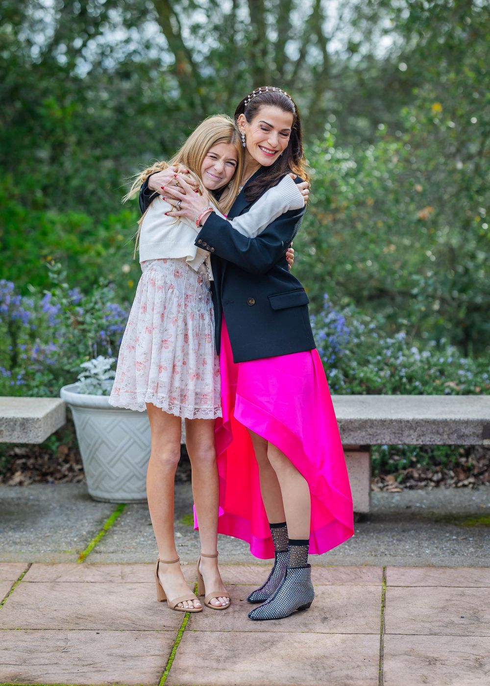 Two smiling girls in dresses embrace on a stone patio beside a bench, with a planter and blooming purple flowers in the garden behind them.