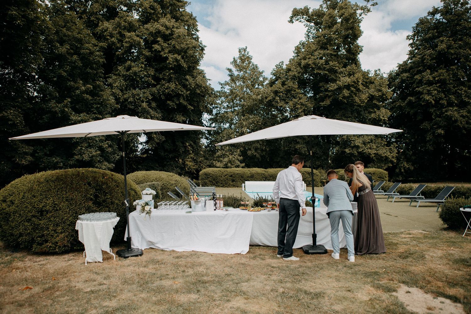 Outdoor wedding reception buffet table with white linens and umbrellas, surrounded by guests and lush green hedges.