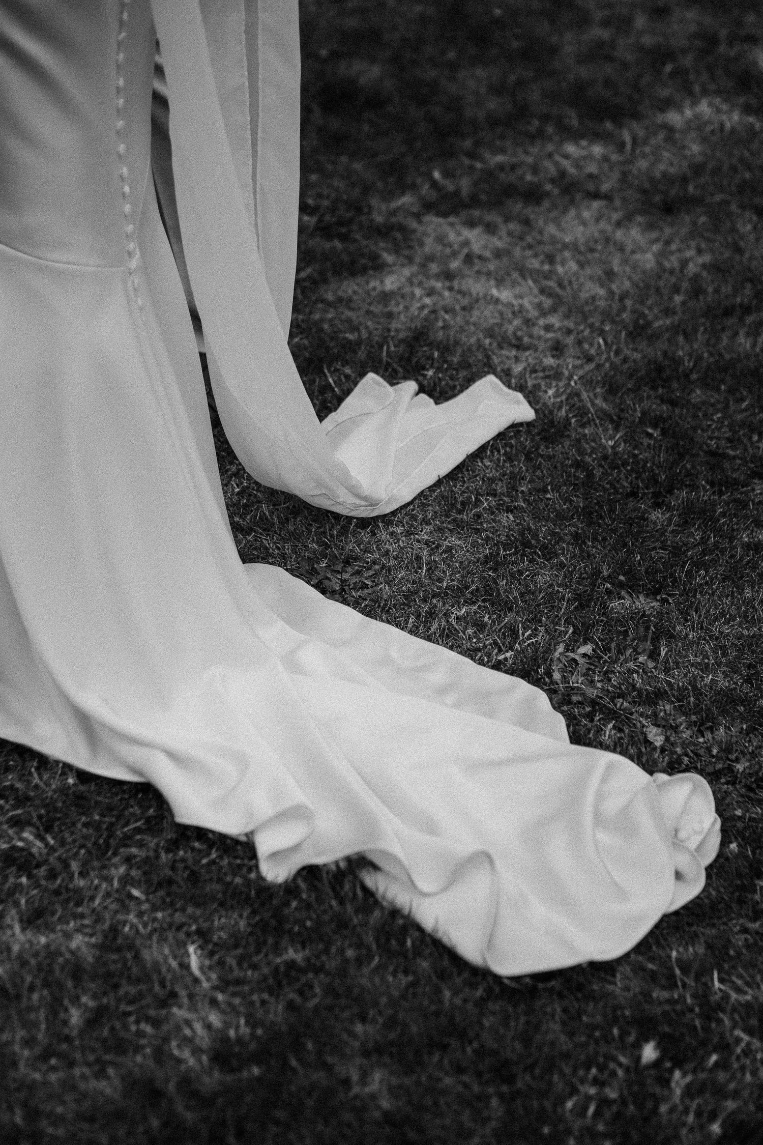 Black and white photo of a wedding dress train flowing gracefully on grass outdoors.
