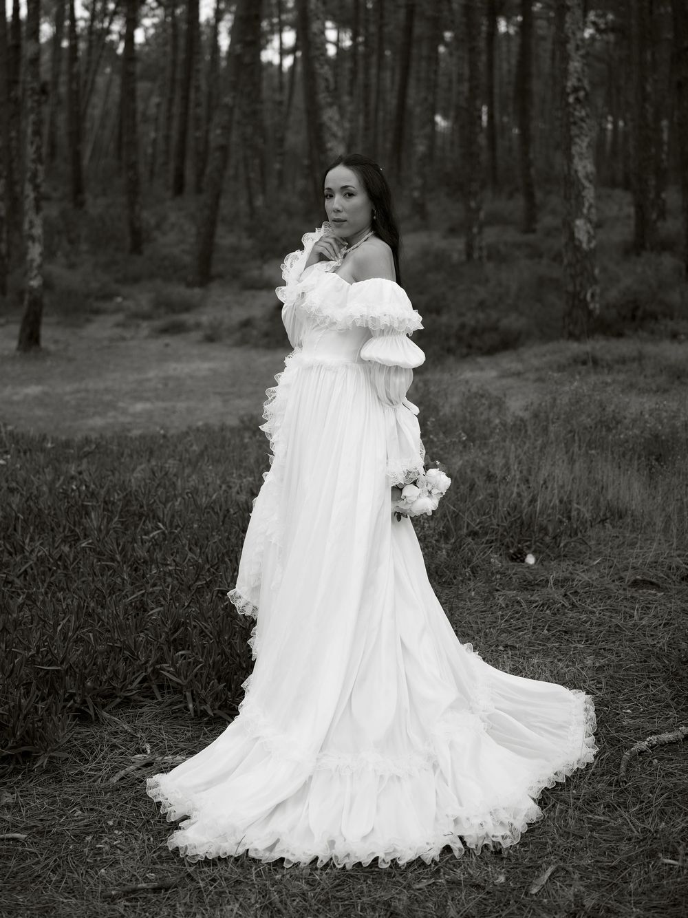 A bride in a flowing white off-shoulder gown with ruffles holds a bouquet in a serene forest setting.