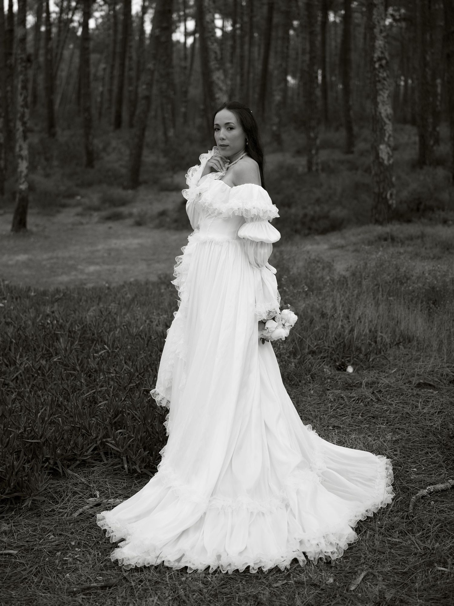 A bride in a flowing white off-shoulder gown with ruffles holds a bouquet in a serene forest setting.