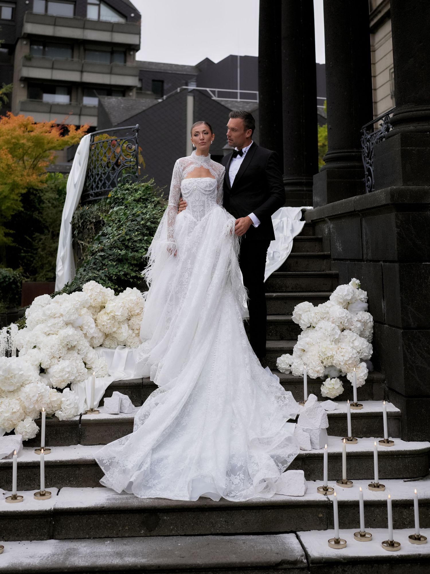 Elegant bride in white lace gown with long train stands with groom in tuxedo on flower-adorned stone steps.