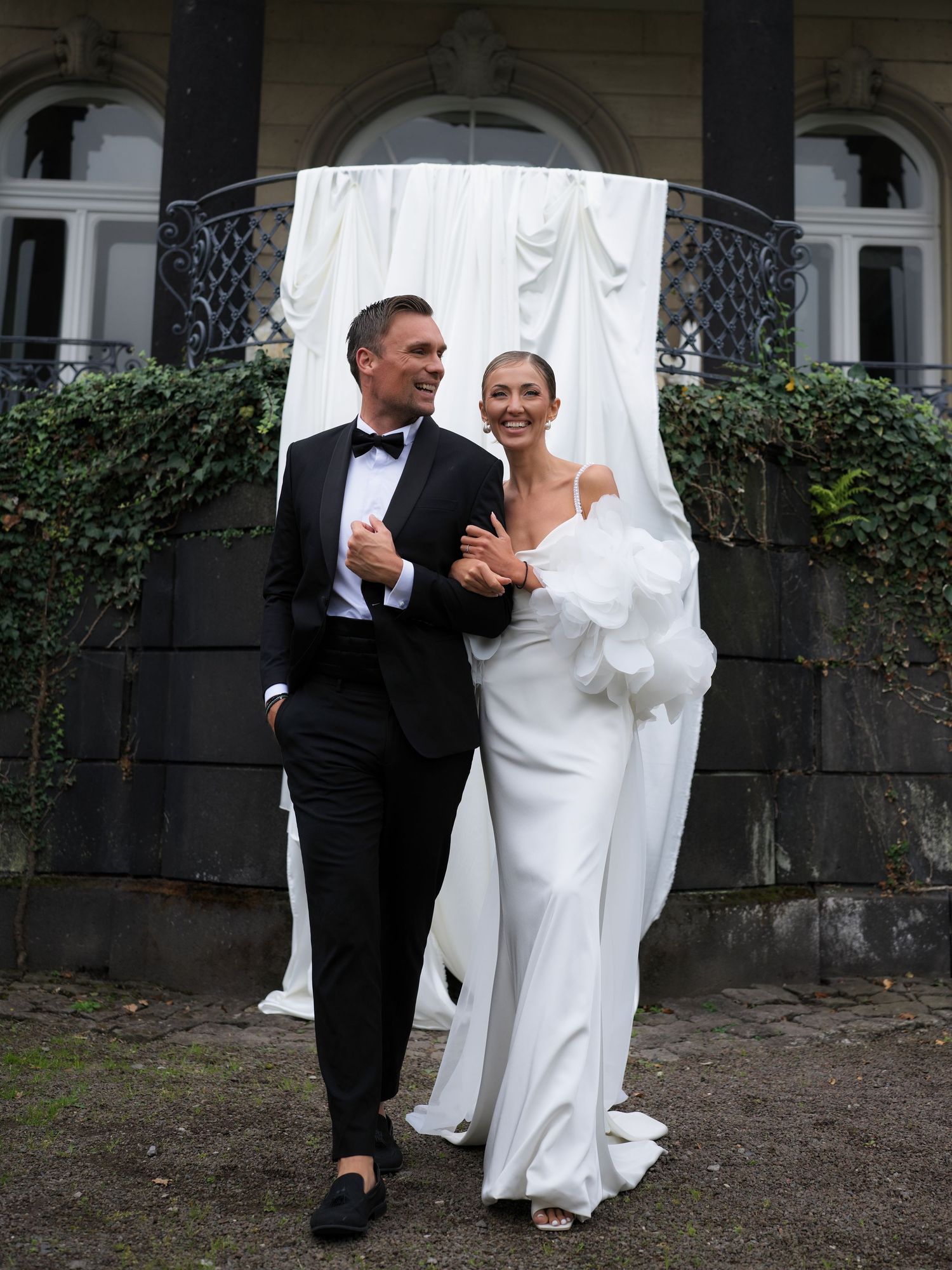 Elegant couple posing in wedding attire, groom in black tuxedo and bride in white gown, before draped backdrop.