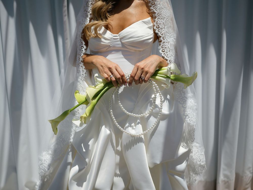 Bride in white wedding gown with lace veil holding calla lily bouquet with pearl accents against white curtain backdrop.