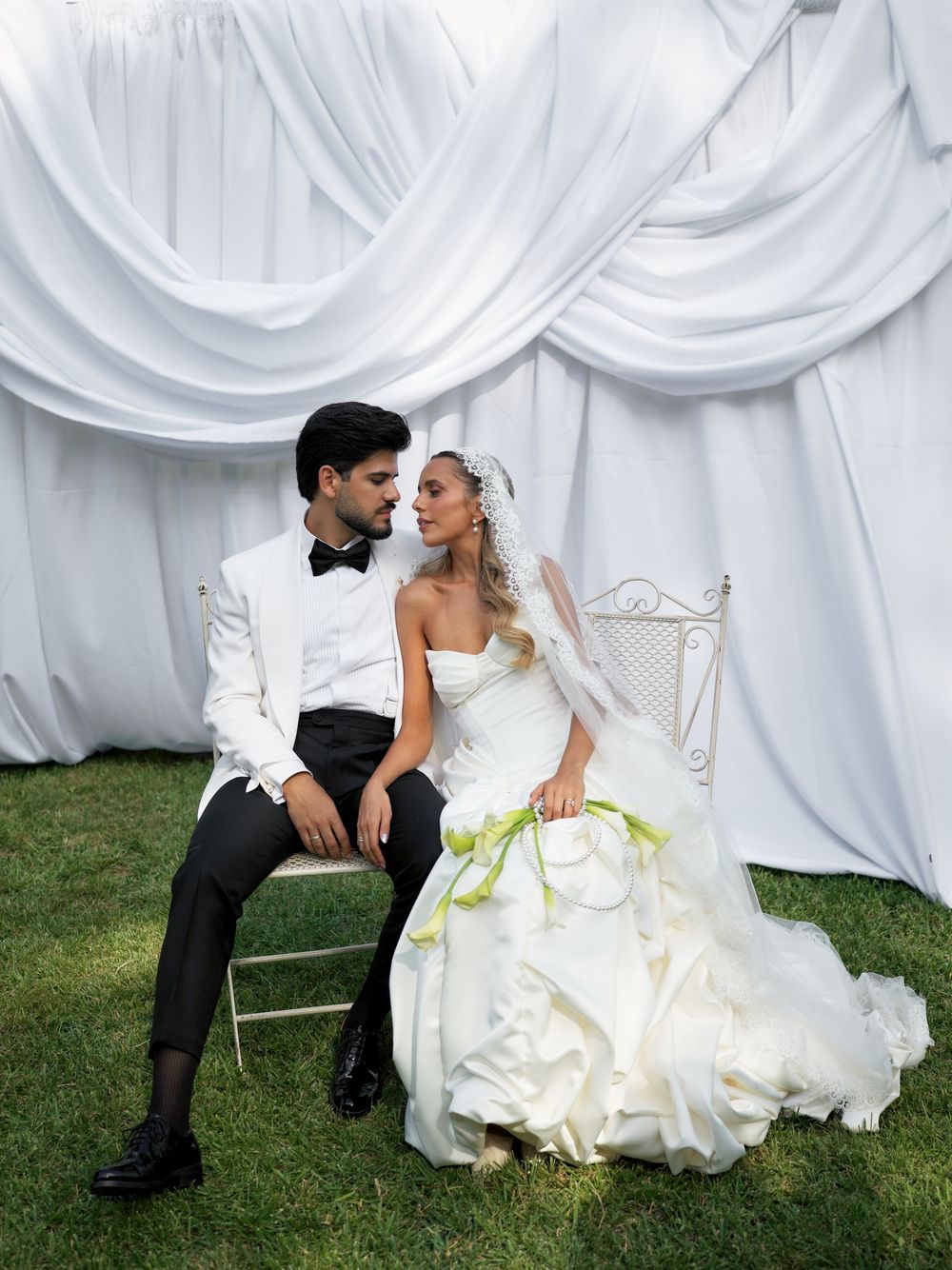A bride in a white gown with green accents and groom in white tuxedo sit together on a chair outdoors.