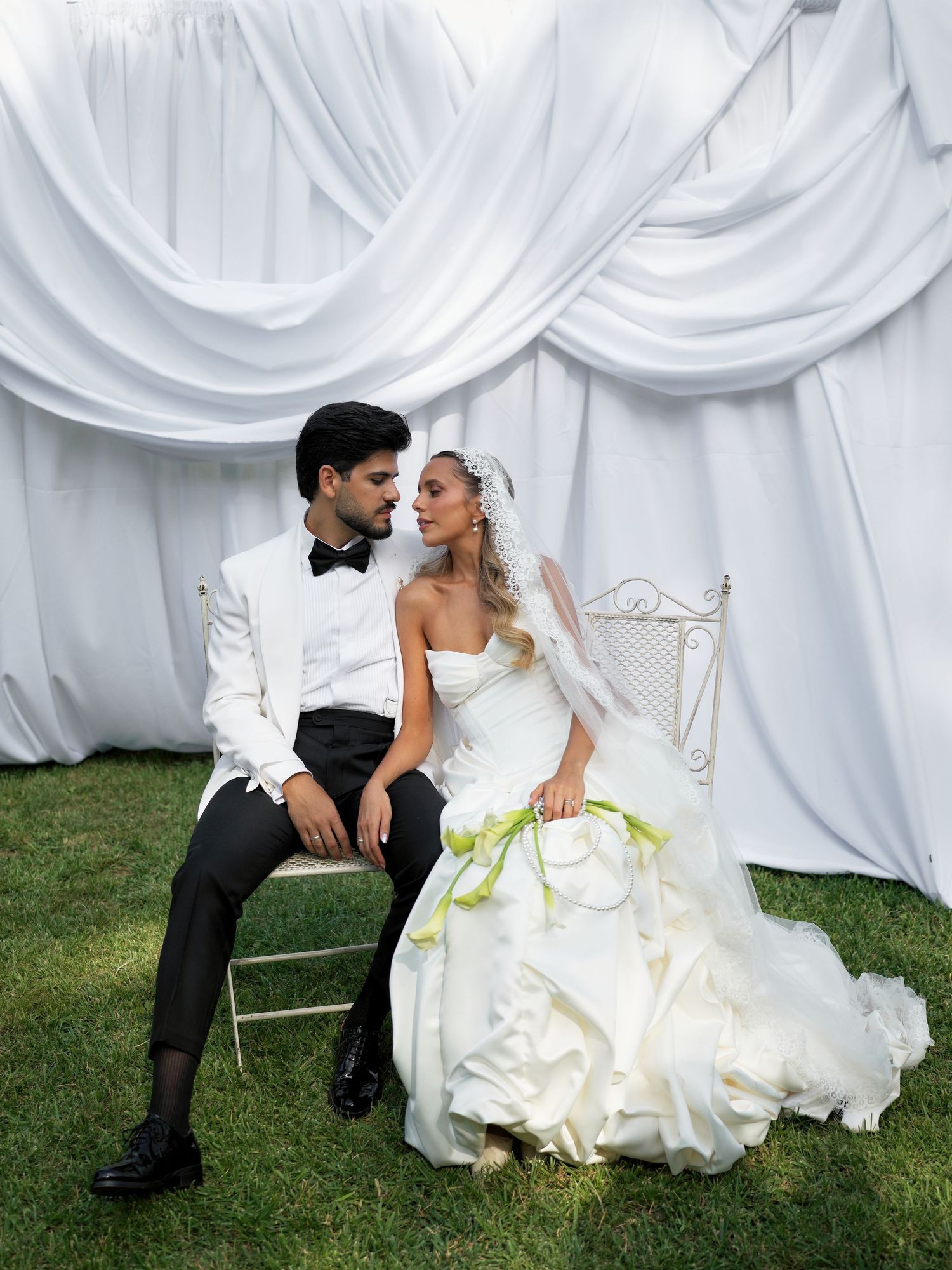 A bride in a white gown with green accents and groom in white tuxedo sit together on a chair outdoors.