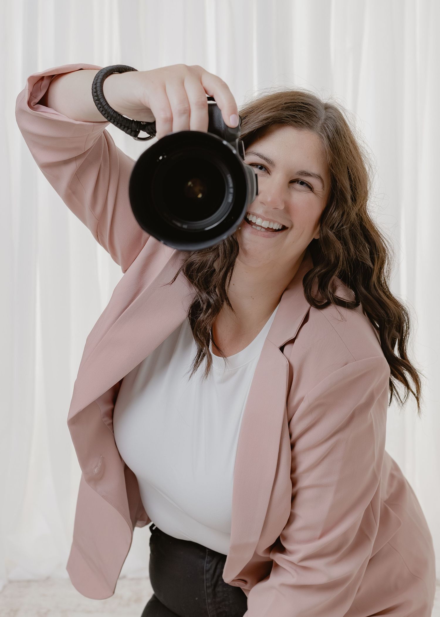 A smiling female photographer in a pink blazer holds a camera up to her face in a bright studio.