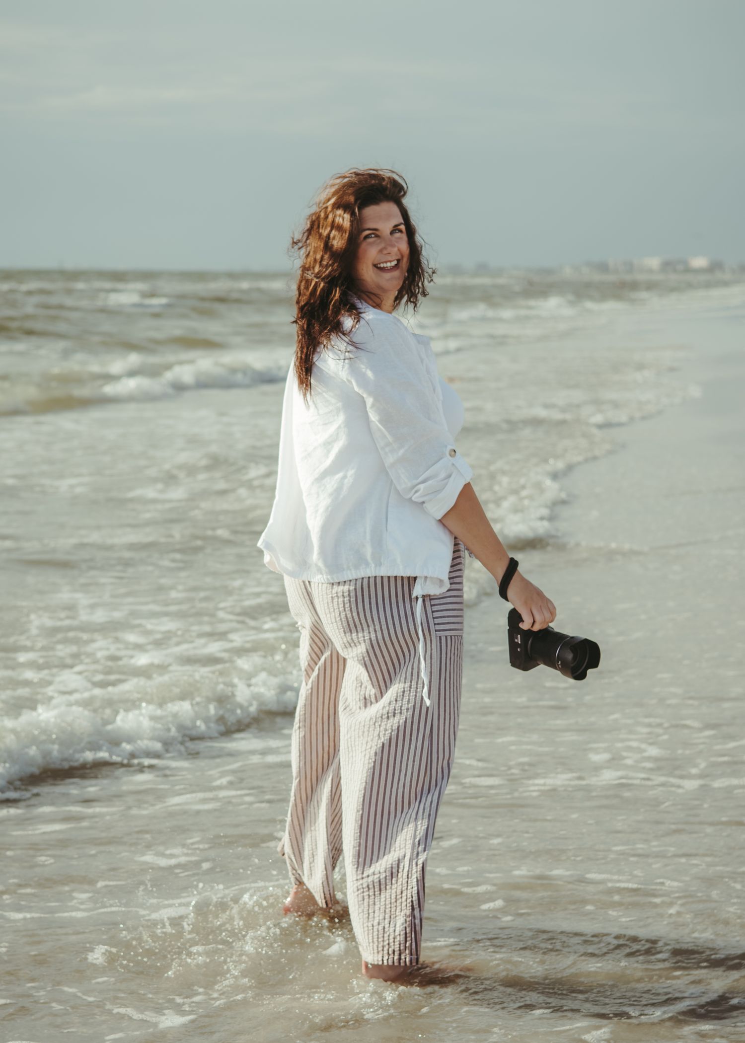 A smiling female photographer in a pink blazer holds a camera up to her face in a bright studio.