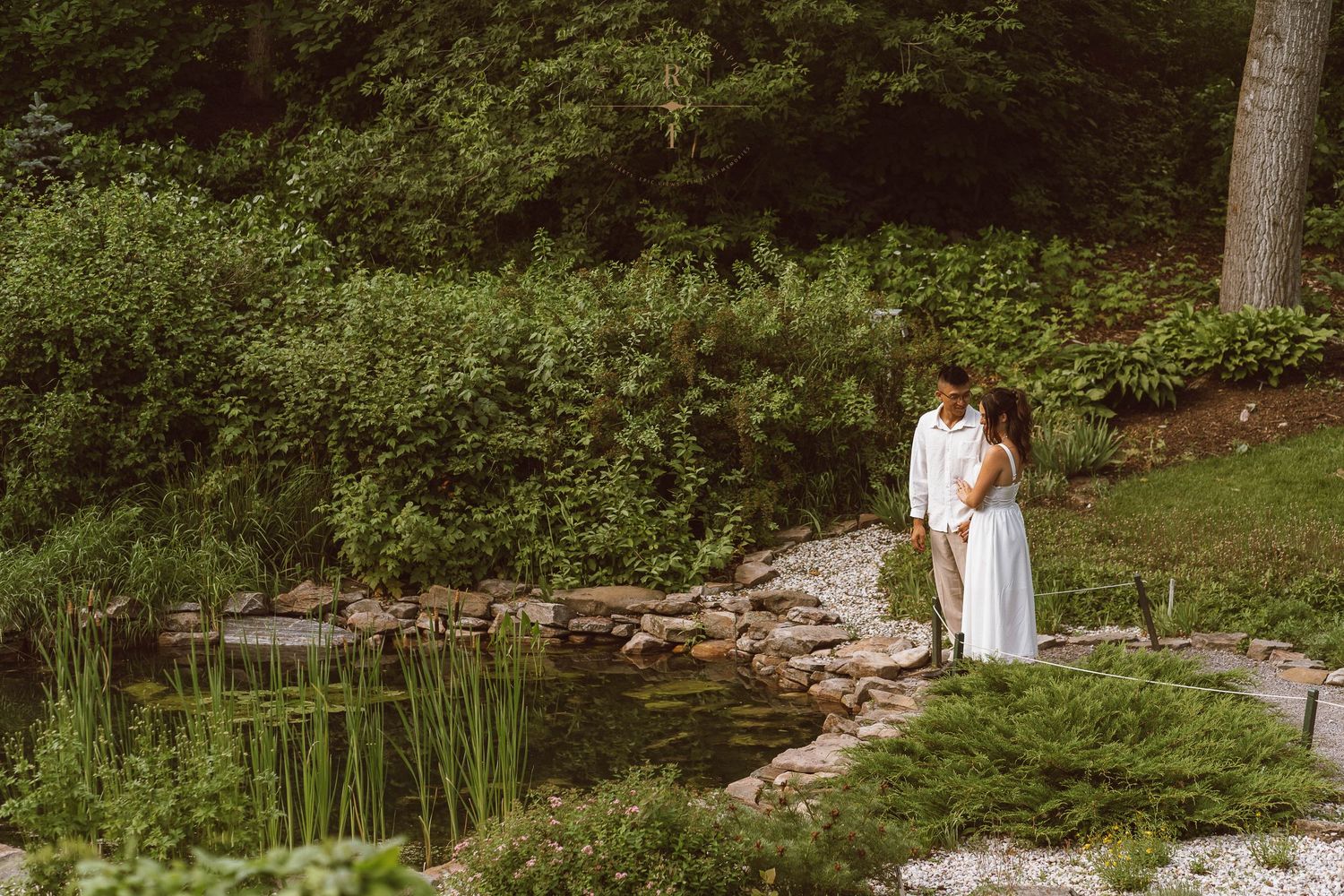 A couple in white attire embraces near a serene garden pond surrounded by lush green trees and plants.