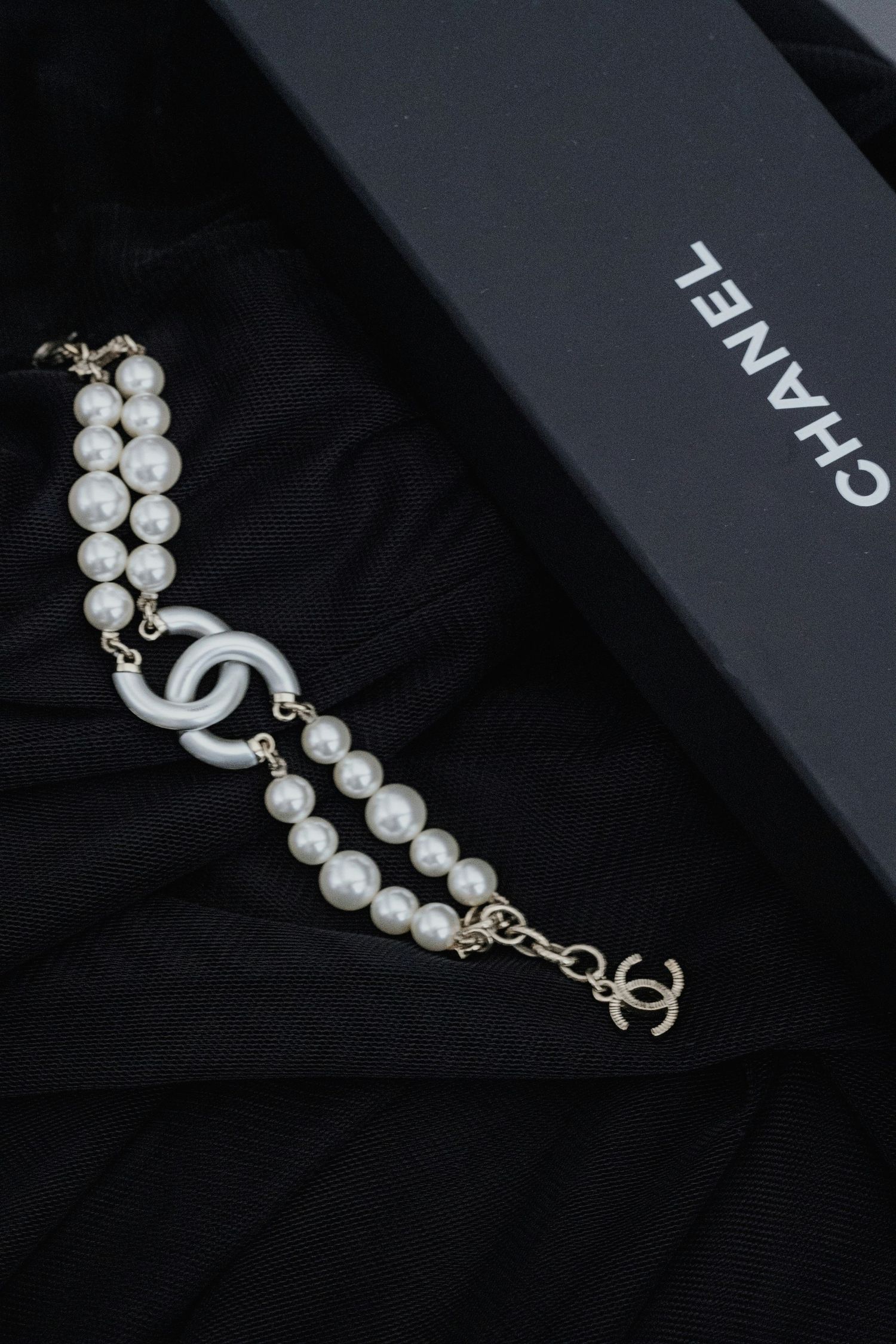 Chanel pearl bracelet with CC logo charm displayed on black fabric beside Chanel gift box.