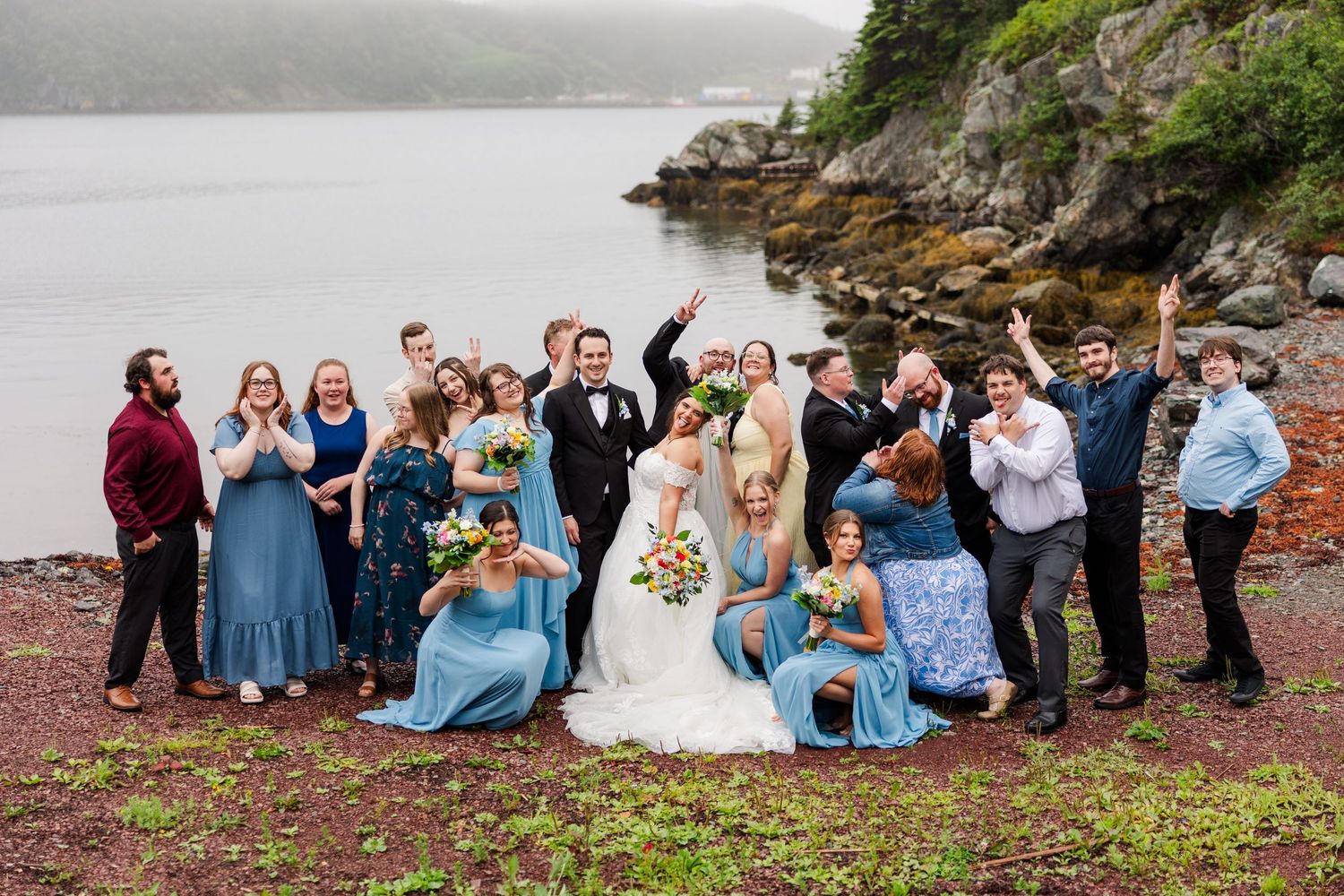 Wedding party celebrating outdoors by a rocky shoreline, bride and groom surrounded by bridesmaids in blue dresses and groomsmen.