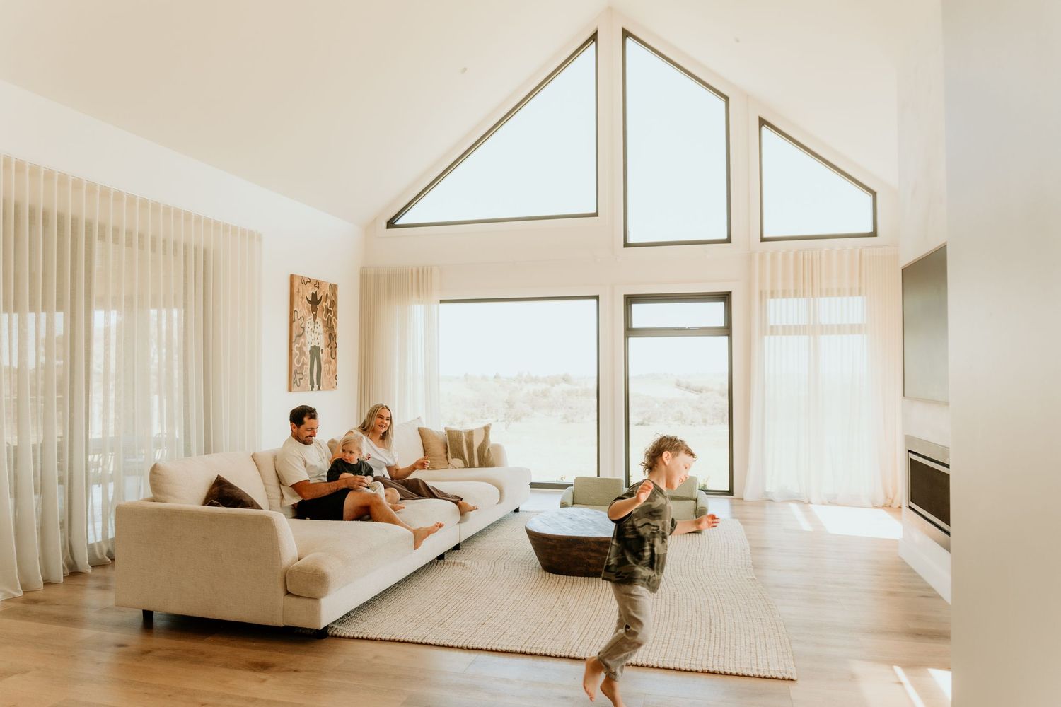 A family relaxes in a bright modern living room with vaulted ceilings and large windows overlooking a scenic landscape.