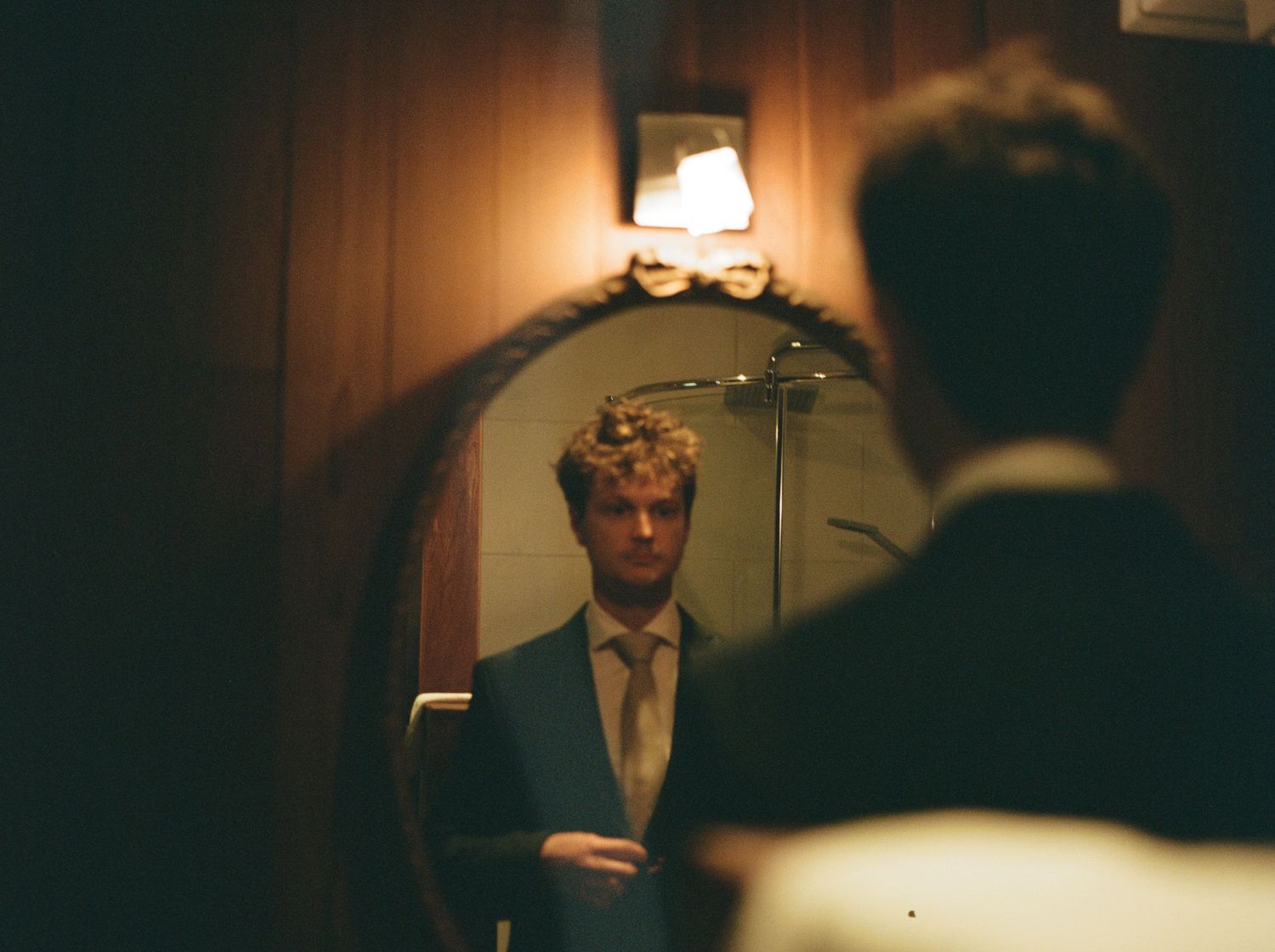 Man in suit reflected in ornate mirror under warm lamp light in dimly lit room.