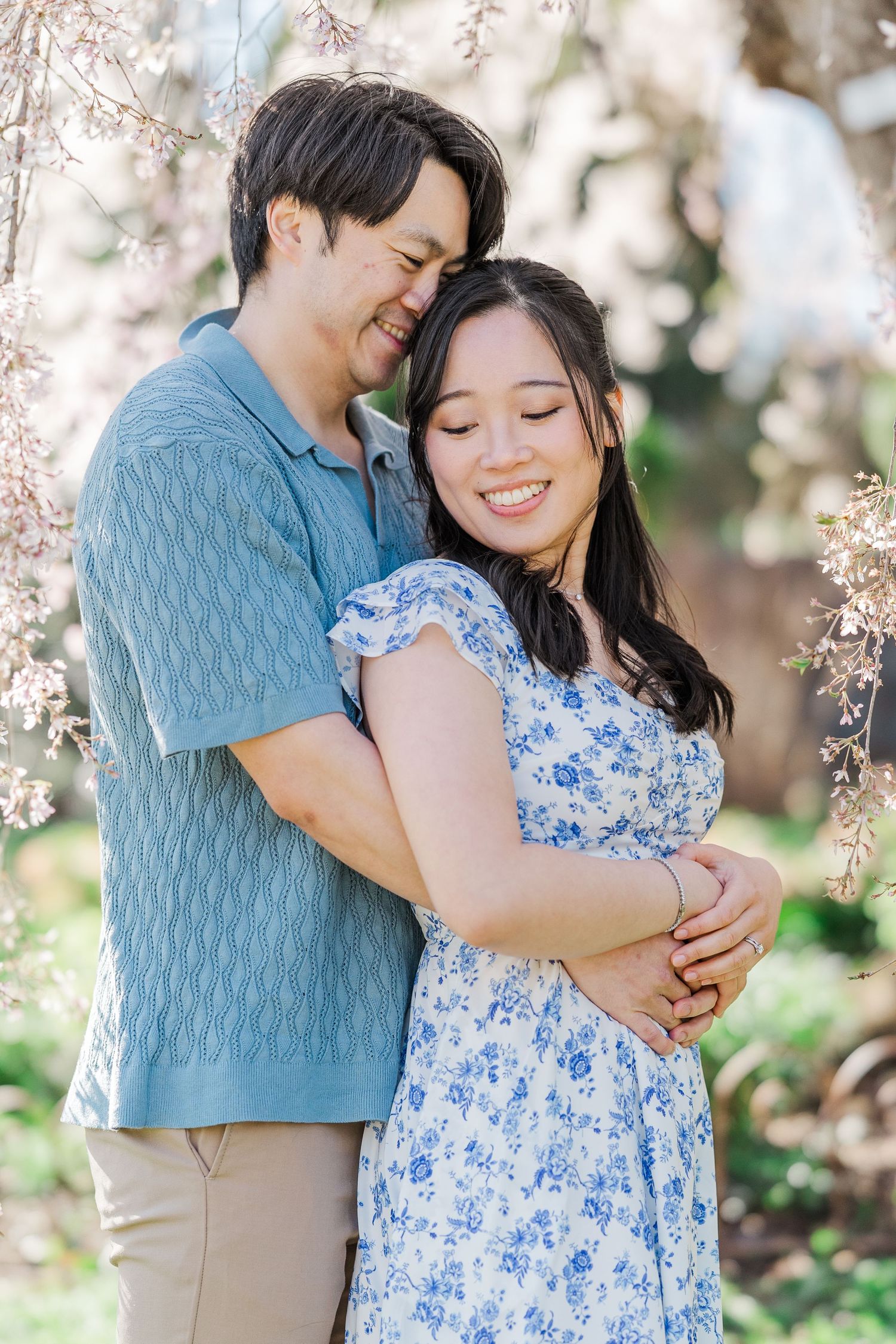 A smiling couple embraces under cherry blossom trees, woman in floral blue dress, man in blue knit polo shirt.