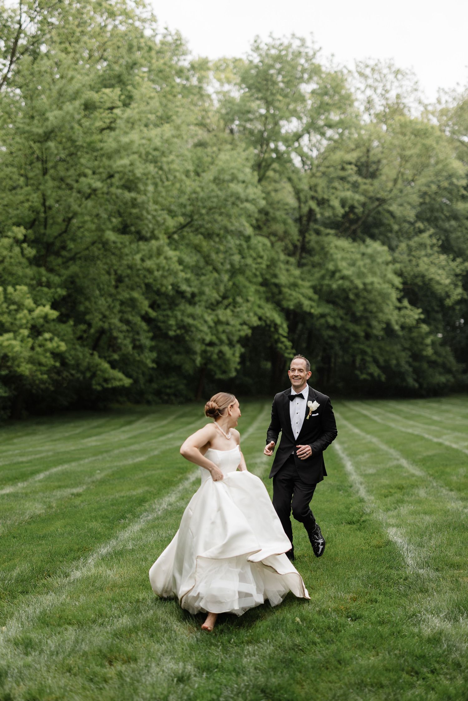 Joyful bride and groom running across lush green lawn during outdoor wedding photo session.