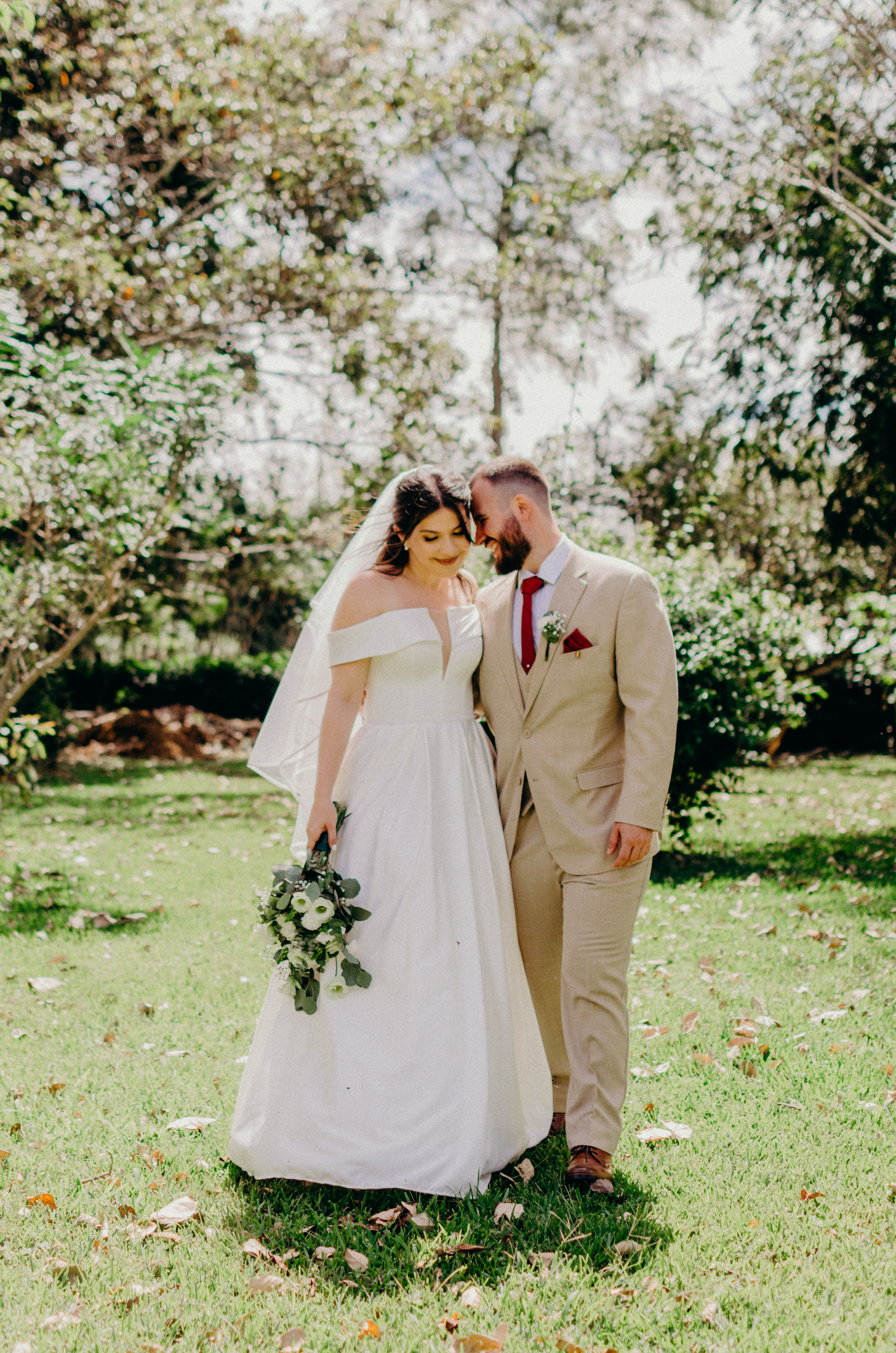 Bride in flowing white gown and groom in tan suit embrace in sunlit garden setting.