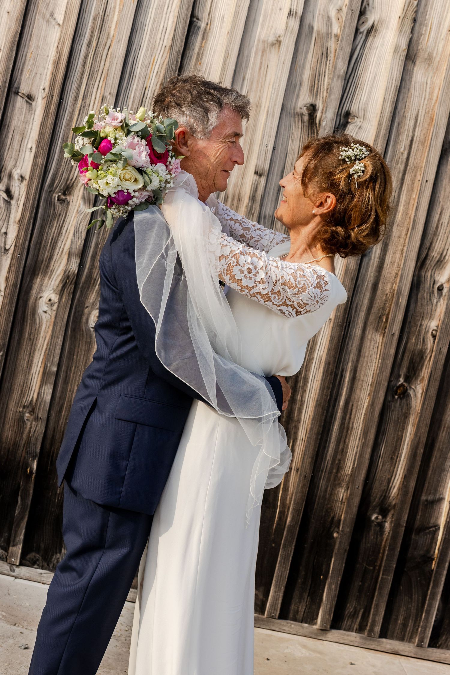Bride and groom embracing romantically in front of rustic wooden wall, bride holding colorful bouquet in white lace gown.
