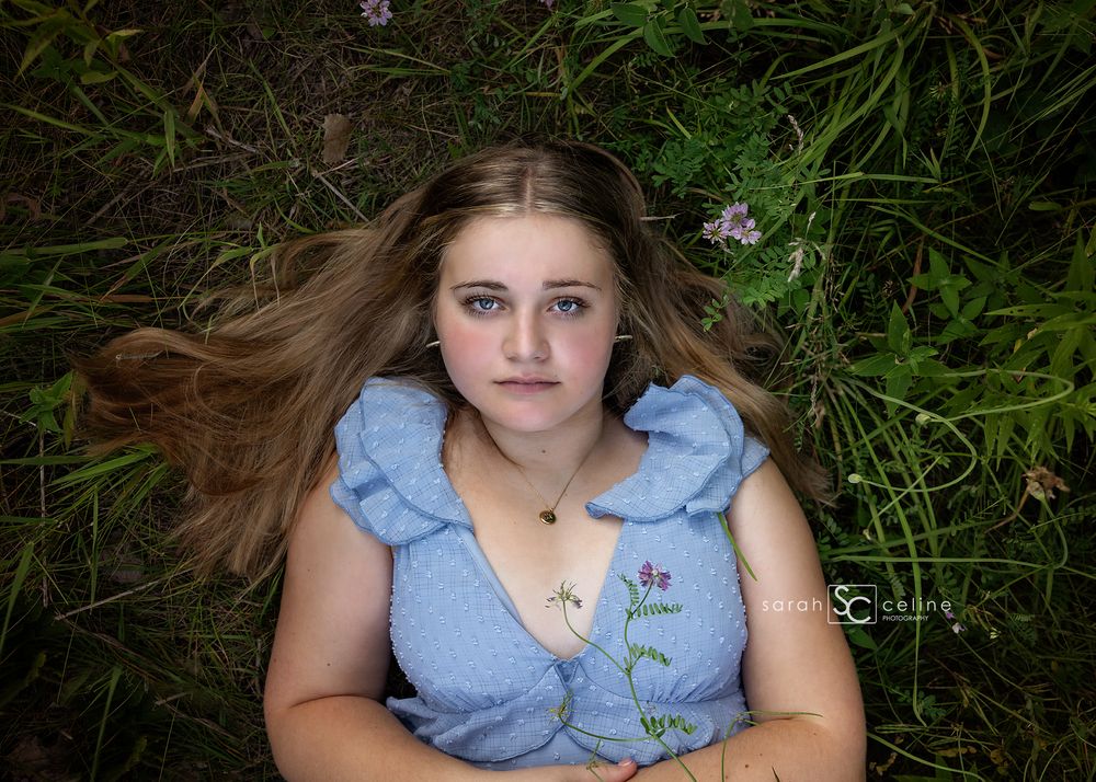 Young girl with long blonde hair lying in green grass wearing a blue floral dress, looking up at camera.