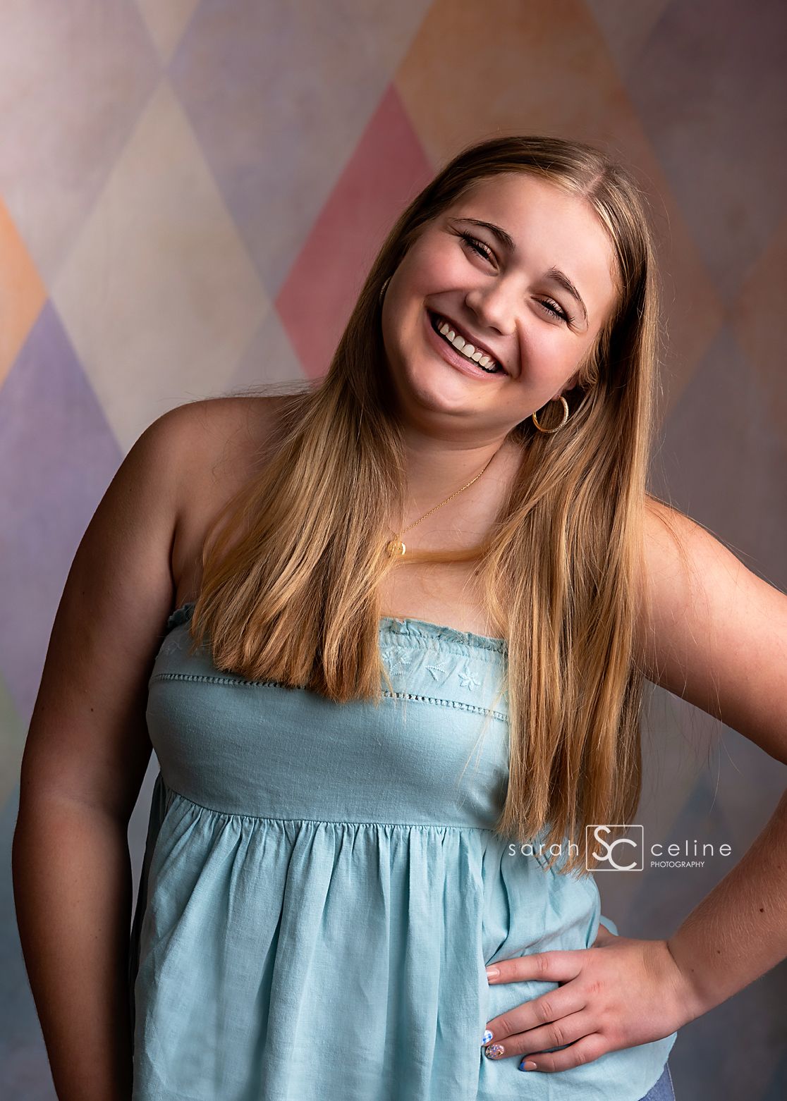 Smiling teenage girl in light blue sundress poses against colorful diamond pattern backdrop in professional portrait.
