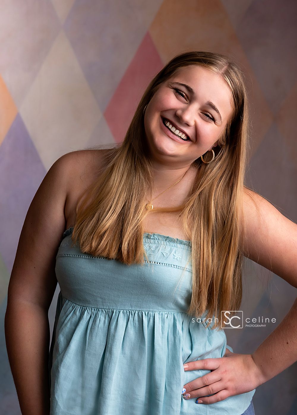 Smiling teenage girl in light blue sundress poses against colorful diamond pattern backdrop in professional portrait.