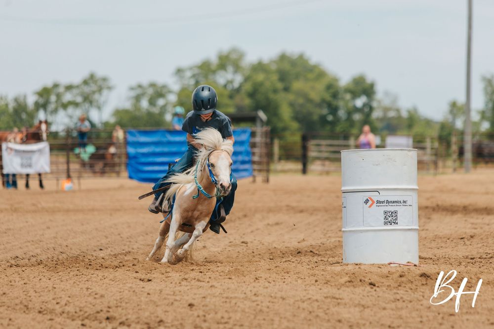 Young rider in helmet races a palomino horse around a barrel in a dirt arena during a barrel racing competition.