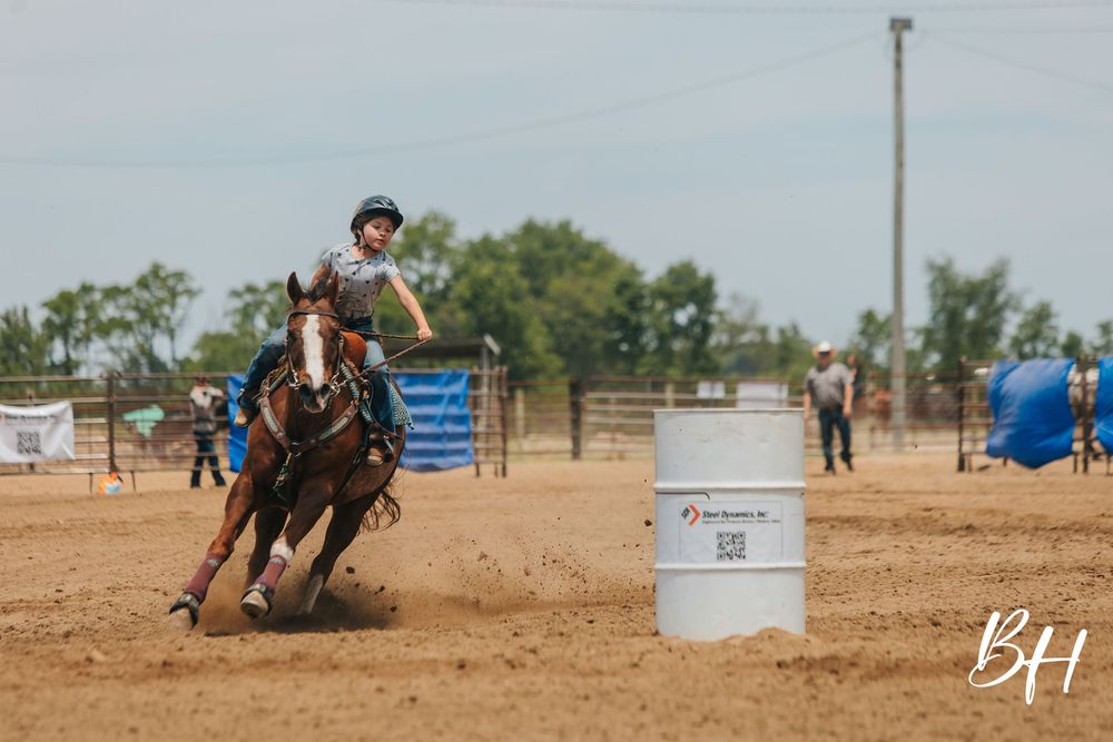 Young rider on horseback competing in barrel racing event at outdoor rodeo arena, navigating around white barrel.