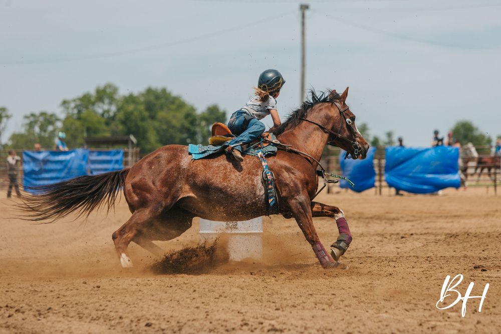 A young rider on a chestnut horse races around a barrel in a dusty arena during a barrel racing competition.