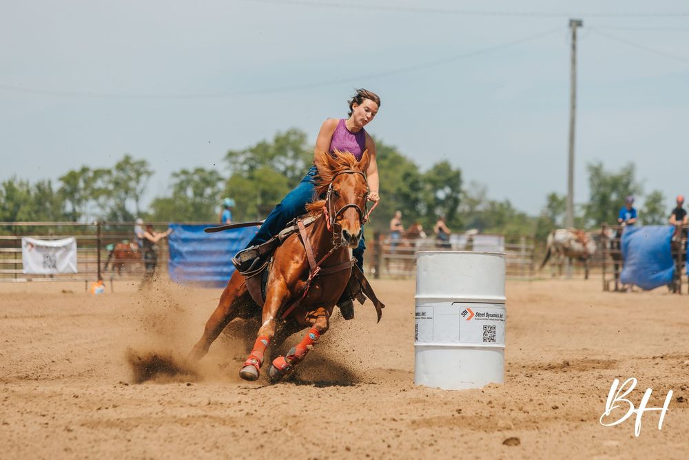 Competitive rider on a chestnut horse makes a sharp turn around a white barrel in a dusty outdoor rodeo arena.