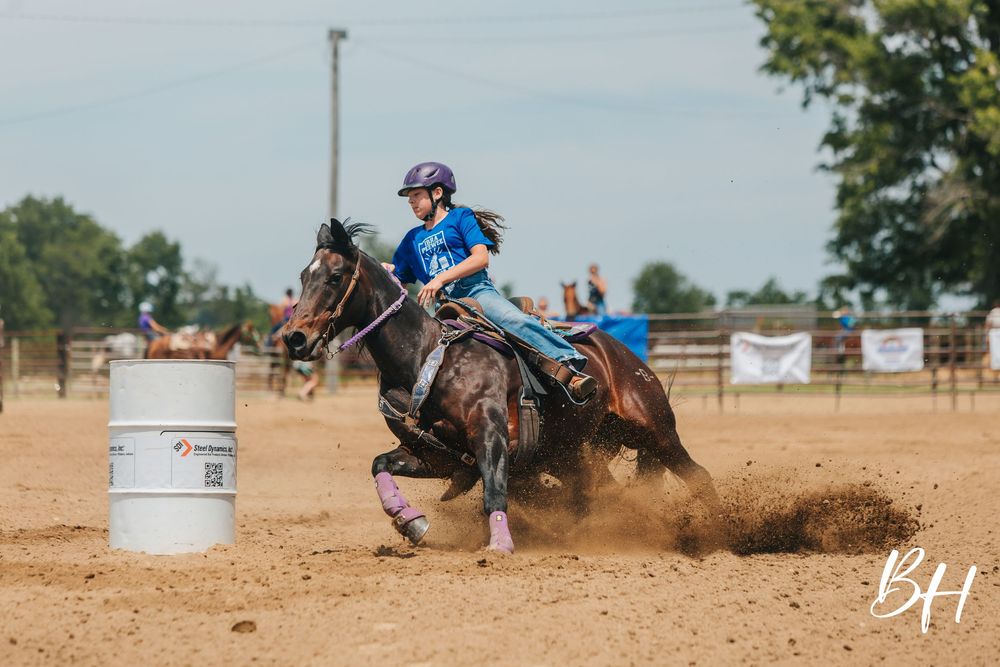 Young girl in purple helmet rides dark horse around barrel in dusty outdoor rodeo arena during barrel racing competition.