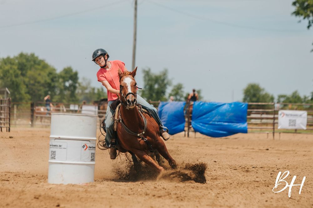 A rider in a pink shirt and helmet races a chestnut horse around a barrel during a rodeo competition on a dirt arena.