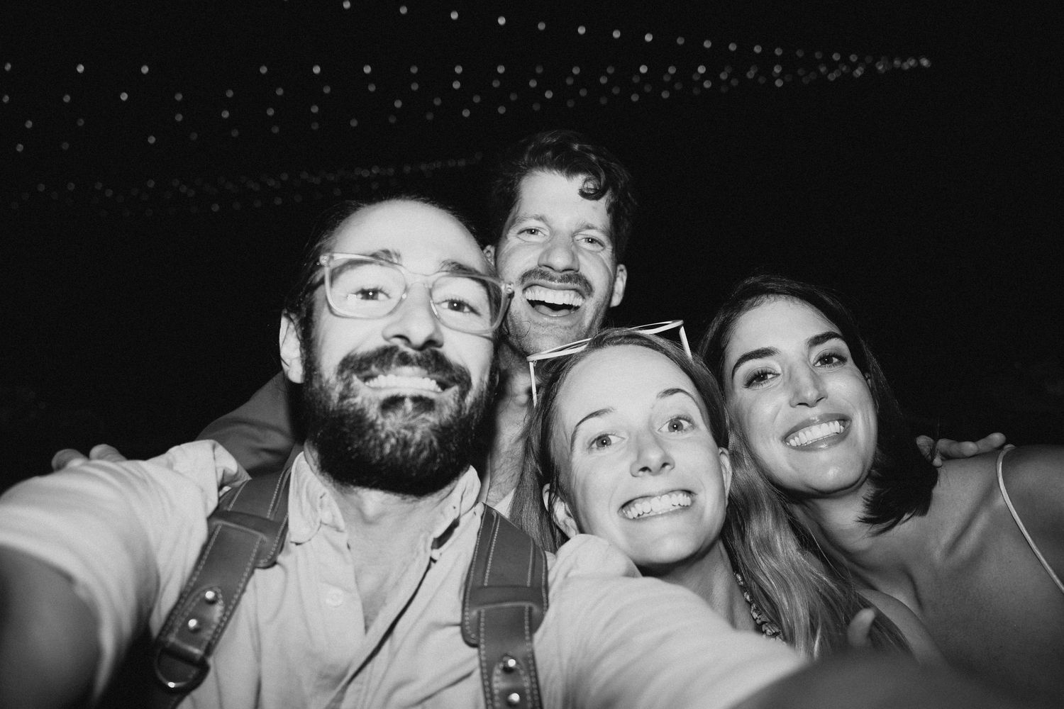 Black and white selfie of four smiling friends at an outdoor night event with string lights in background.
