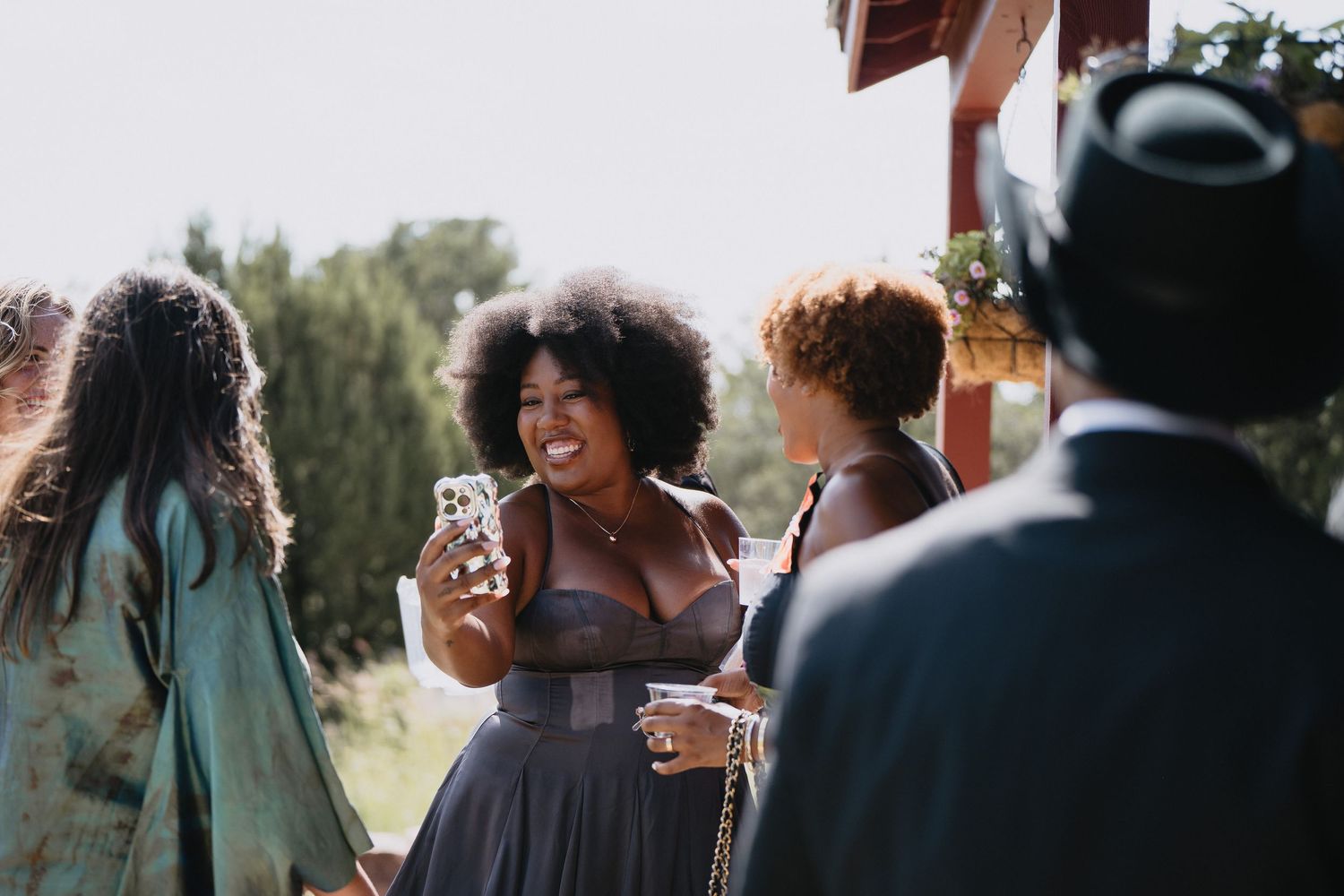 Joyful woman with natural afro hair smiling and holding phone at outdoor social gathering with friends.