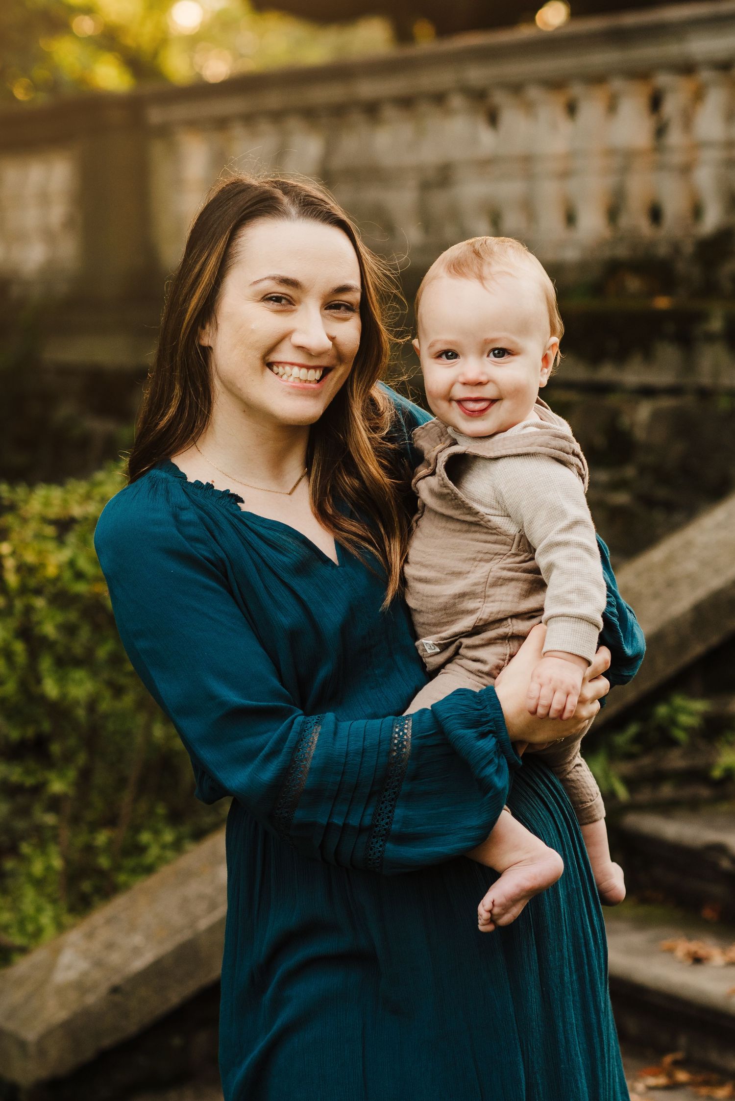 A smiling woman in a teal dress holds a happy baby outdoors near stone steps with lush greenery.