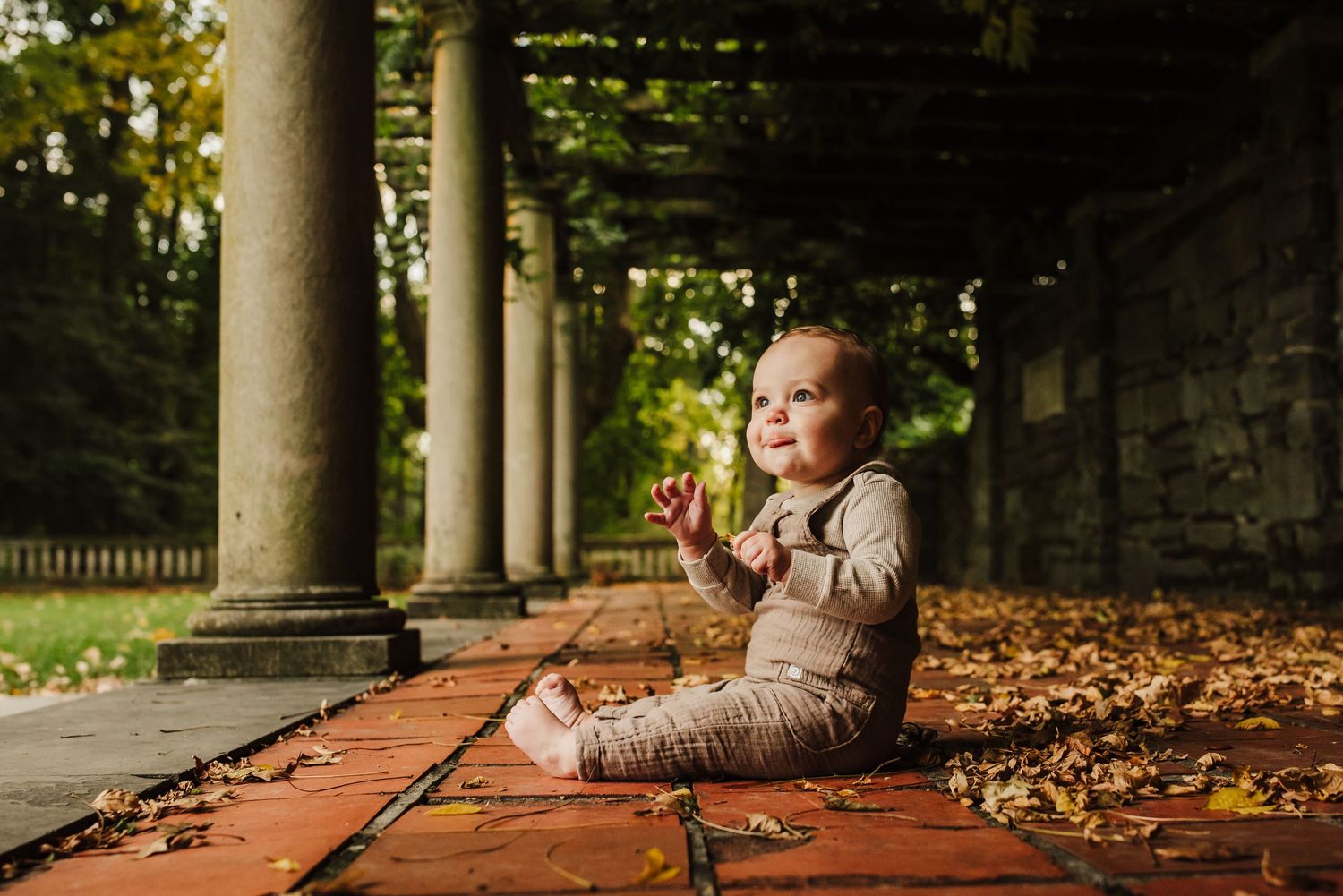 Baby sitting on brick floor among autumn leaves under stone columns in a lush garden setting.