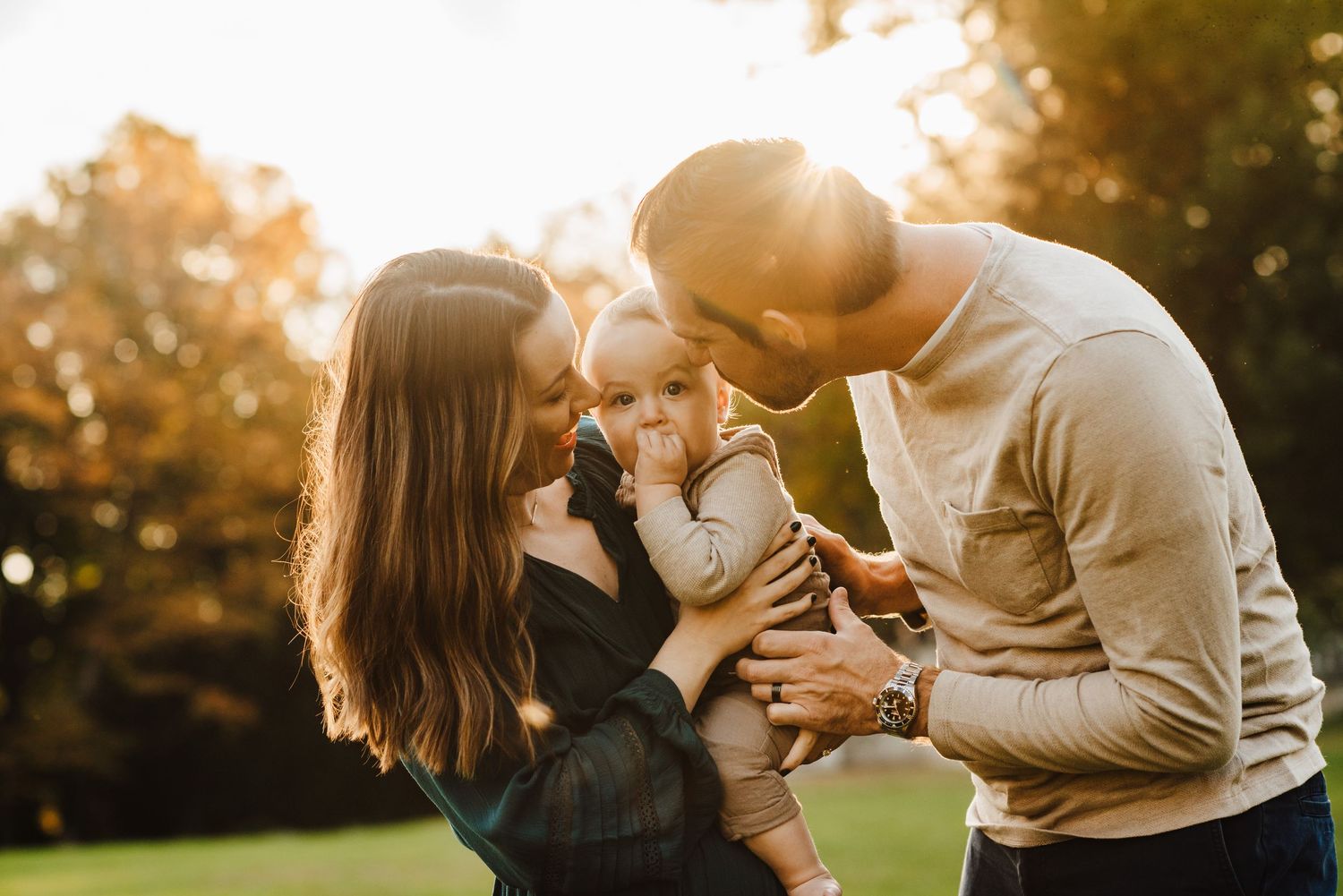 Young family with baby enjoying golden hour sunlight in autumn park, parents smiling at infant outdoors.