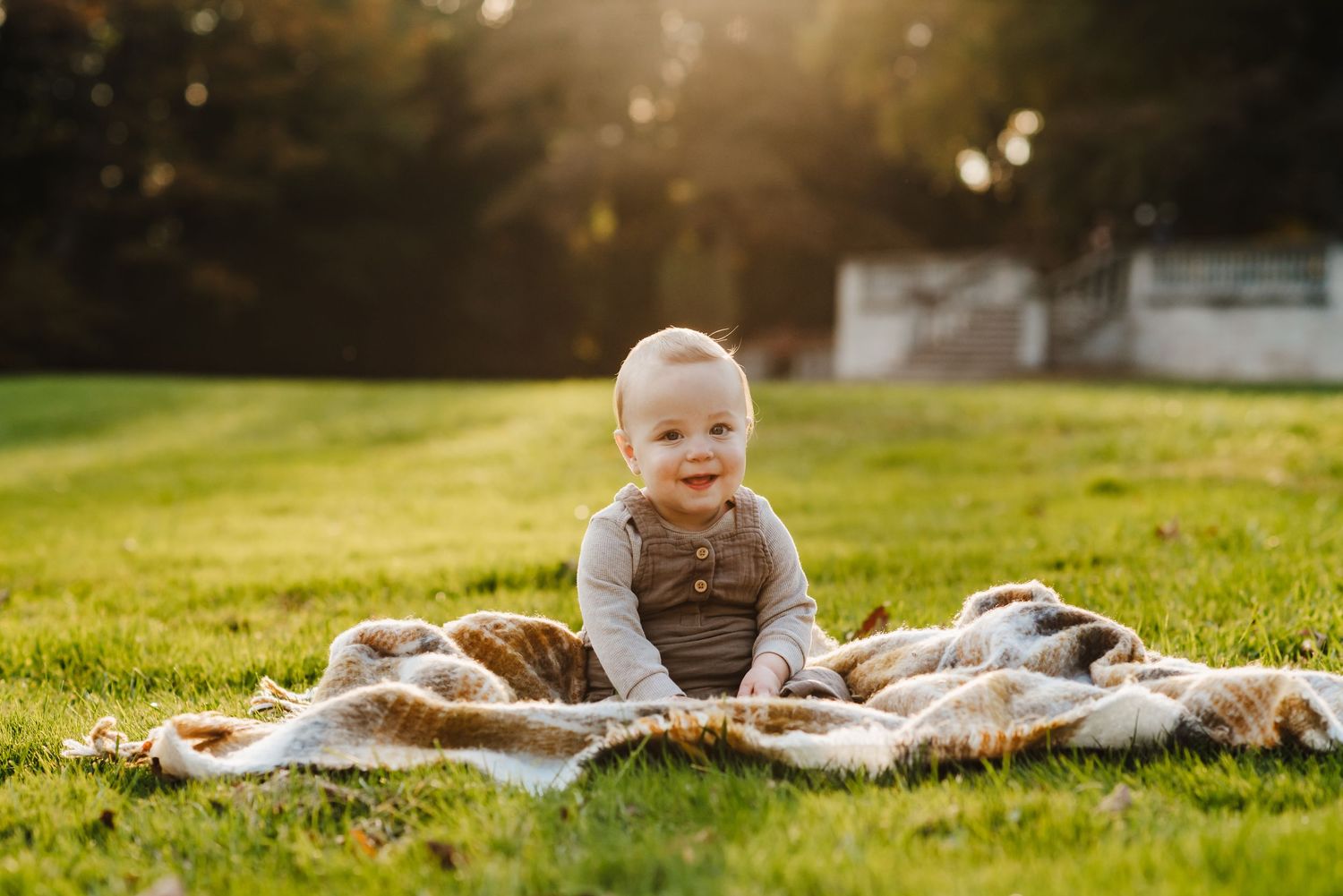 A happy baby sitting on a plaid blanket in a sunny green park, smiling in warm golden hour light.