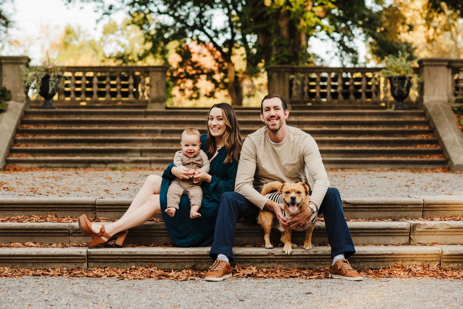 Family portrait with parents, baby, and golden retriever sitting on stone steps surrounded by autumn foliage.