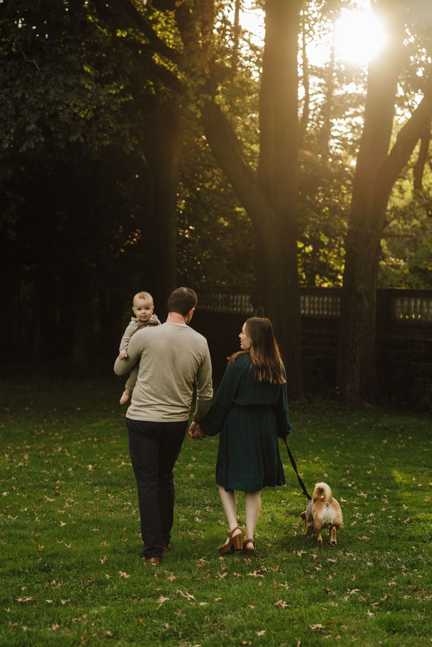 A family walks through a sunlit park at golden hour, with dad carrying a baby, mom holding a small dog on a leash.