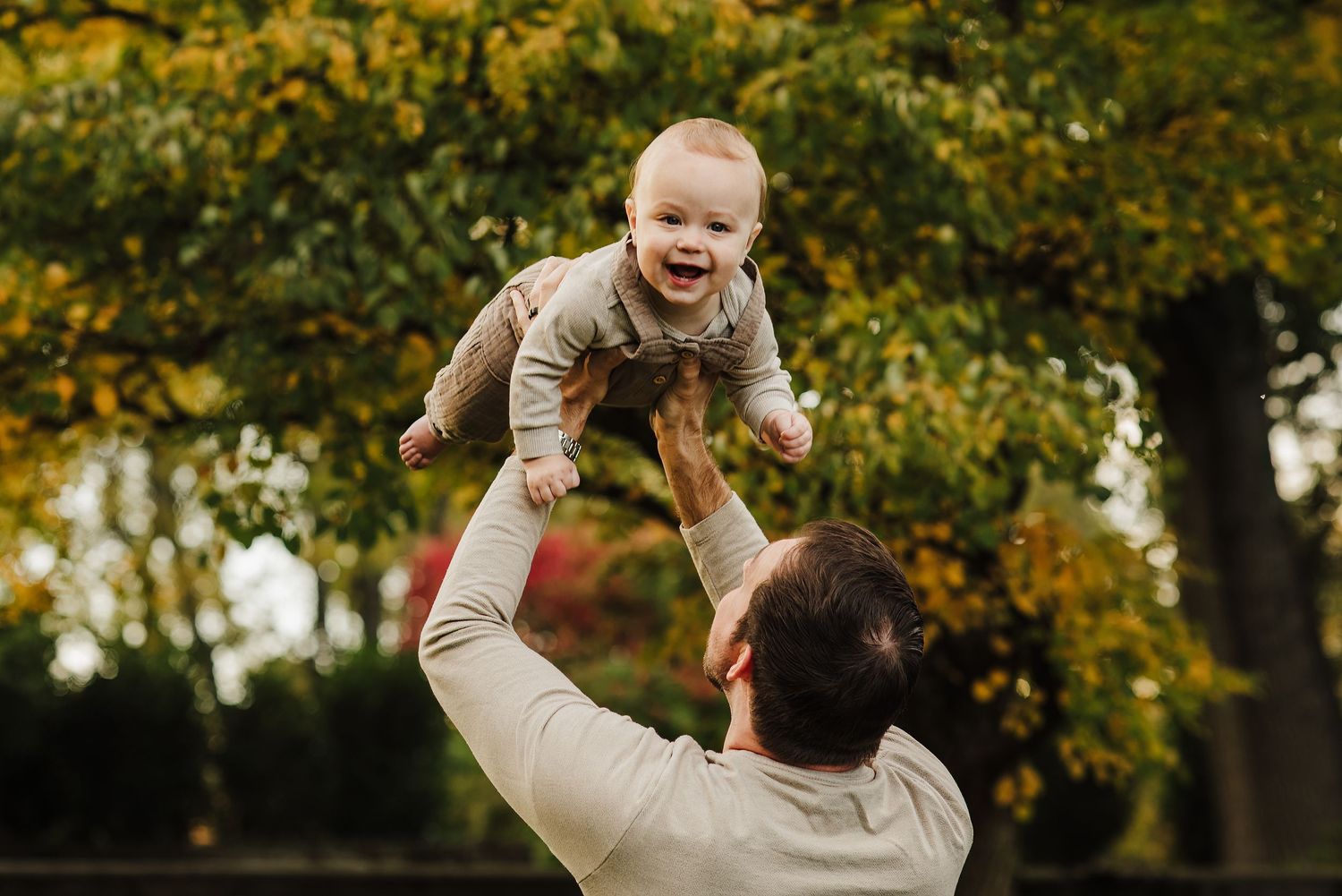 Father joyfully tosses laughing baby in the air outdoors, surrounded by colorful autumn foliage in a park.