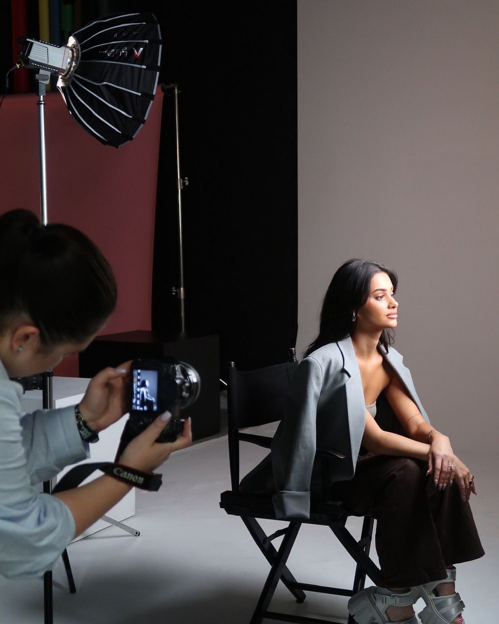 Photographer capturing model during studio photoshoot with lighting equipment visible.