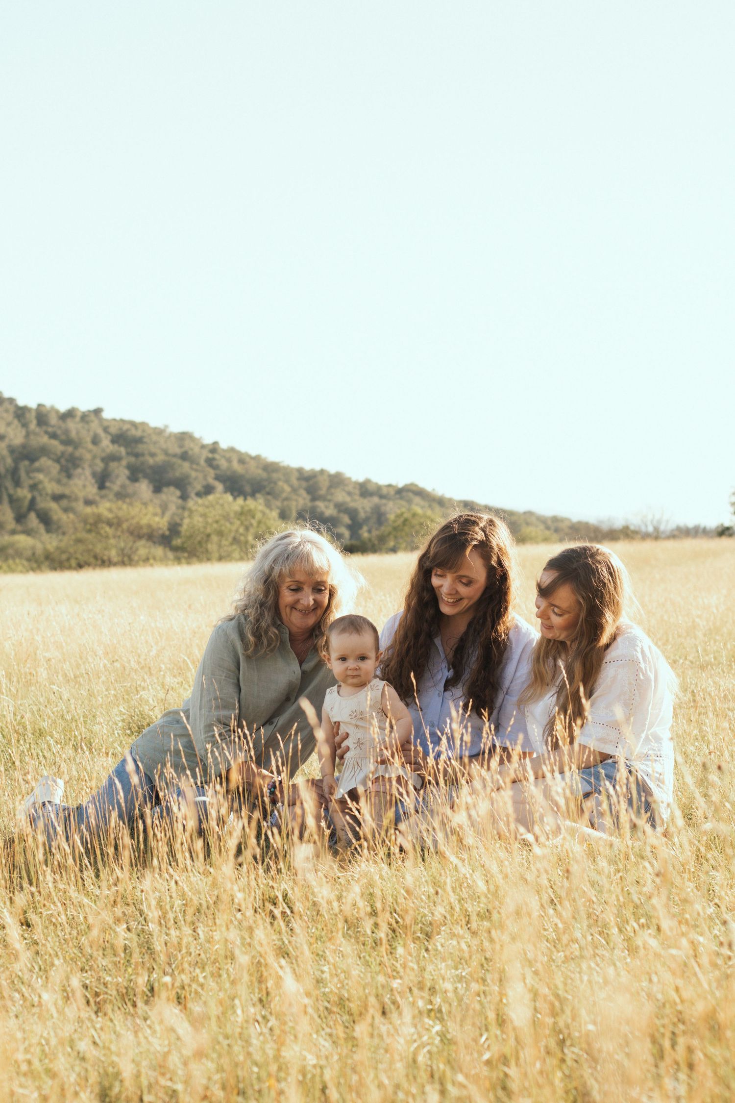 Four generations of women sitting together in a golden field, smiling at a baby on a sunny day.