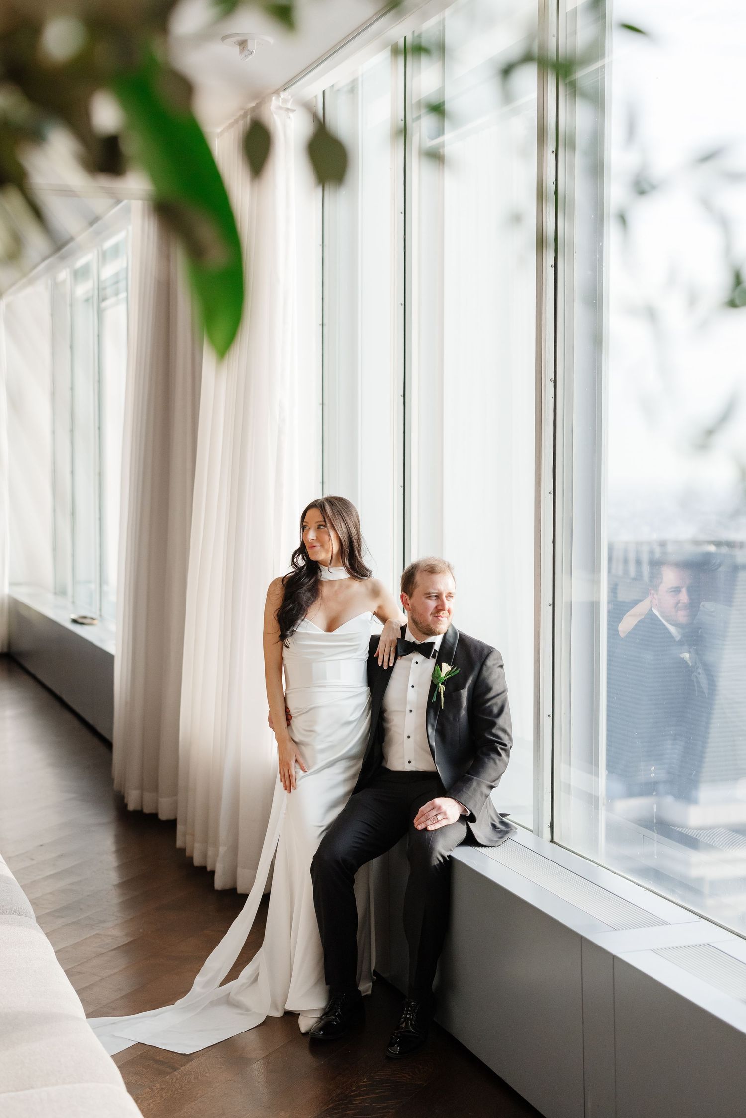 Bride in white gown and groom in black tuxedo pose by floor-to-ceiling windows on their wedding day.