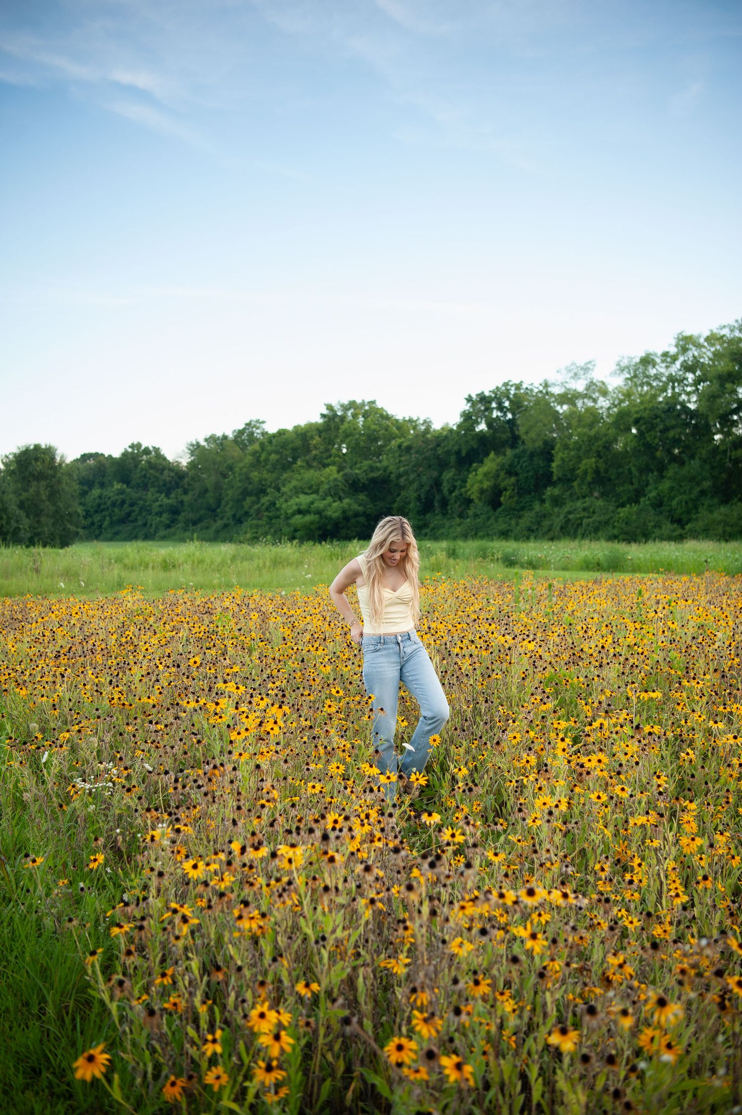 Blonde woman in yellow top and jeans standing in a vibrant field of yellow wildflowers with green trees in background.