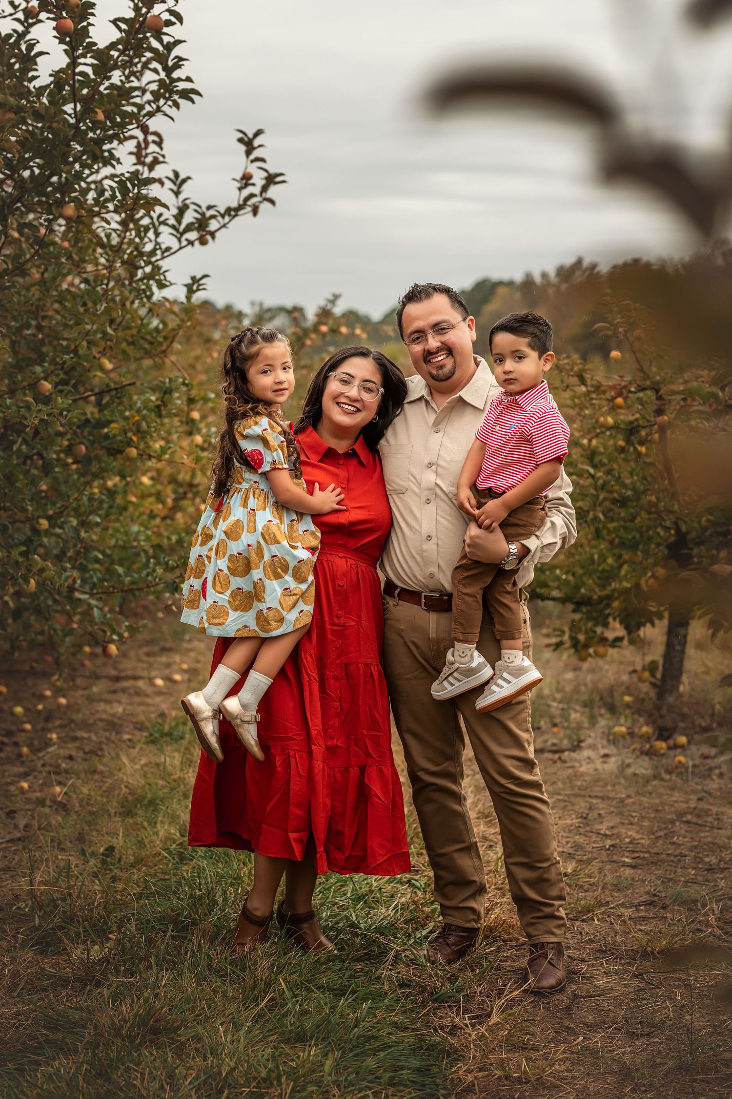 Family of four posing in an apple orchard, parents holding young children, woman in red dress, fall setting.