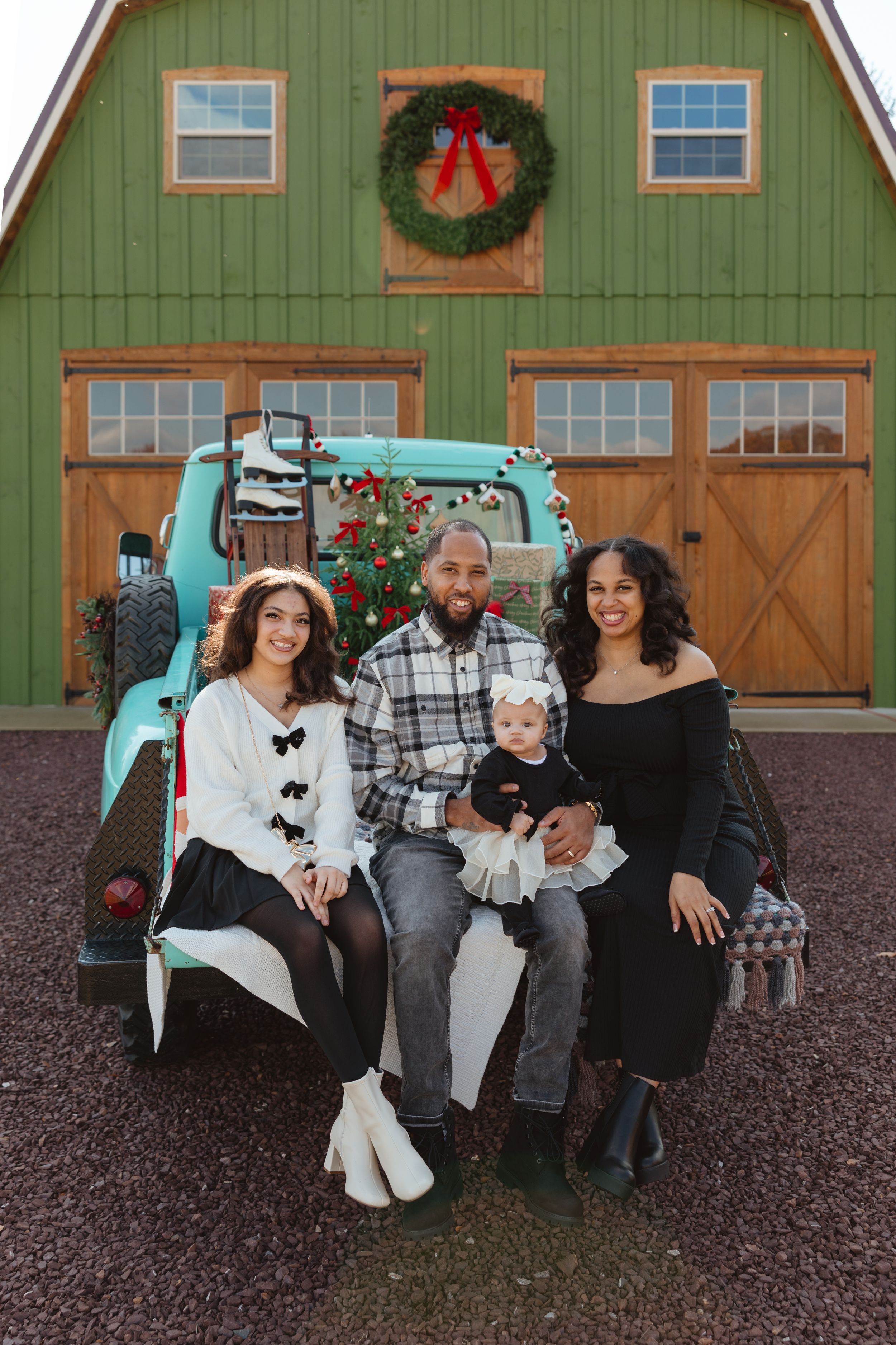 Family of four poses on vintage teal truck bed before green barn with Christmas wreath for holiday portrait.